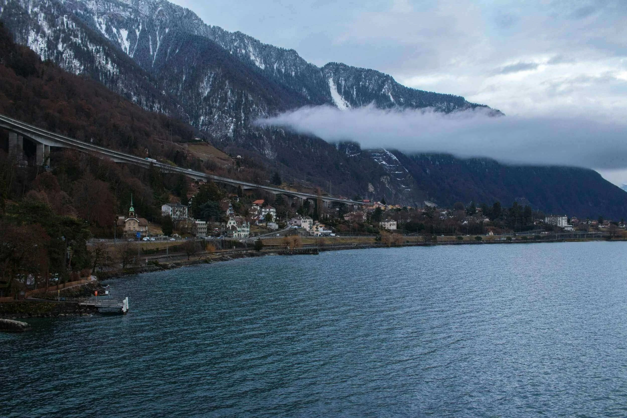 Vue panoramique de Montreuil, avec un lac calme entouré de montagnes enneigées et une couche de brume ajoutant une atmosphère mystérieuse. Idéal pour découvrir notre agence et ses services d’essayage dans cette ville pittoresque et naturelle.