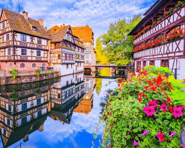 Vue pittoresque du quai des Bateliers à Strasbourg, avec des maisons à colombages reflétées dans l’eau et des fleurs colorées en bordure. Idéal pour découvrir comment devenir mannequin d’essayage à Strasbourg, une ville aux charmes alsaciens.