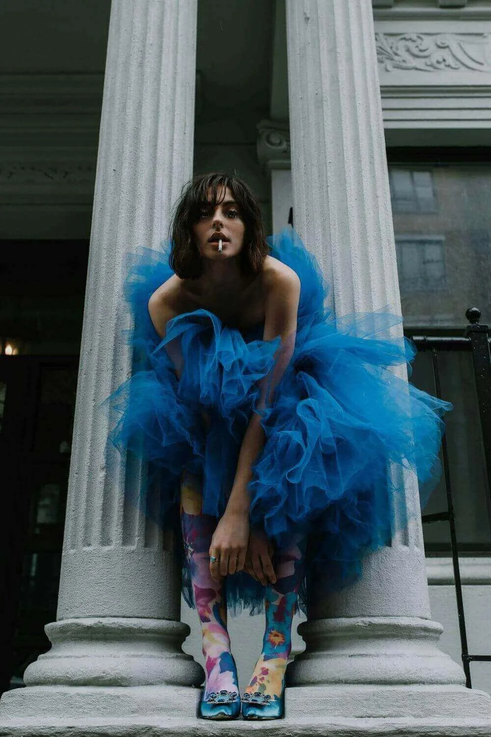 Image d’une mannequin en tenue créative : robe en tulle bleu, collants fleuris et chaussures assorties. Pose audacieuse avec une cigarette, entre deux colonnes classiques. Ambiance mode éditoriale moderne et élégante.