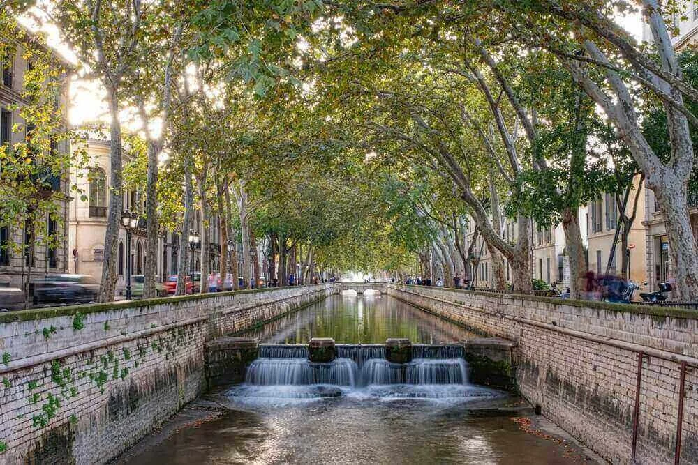Vue paisible du canal de la Fontaine du Gard à Nîmes, bordé d’arbres et avec une chute d’eau. Idéal pour découvrir comment devenir mannequin d’essayage à Nîmes, une ville où l’architecture romaine côtoie la nature.