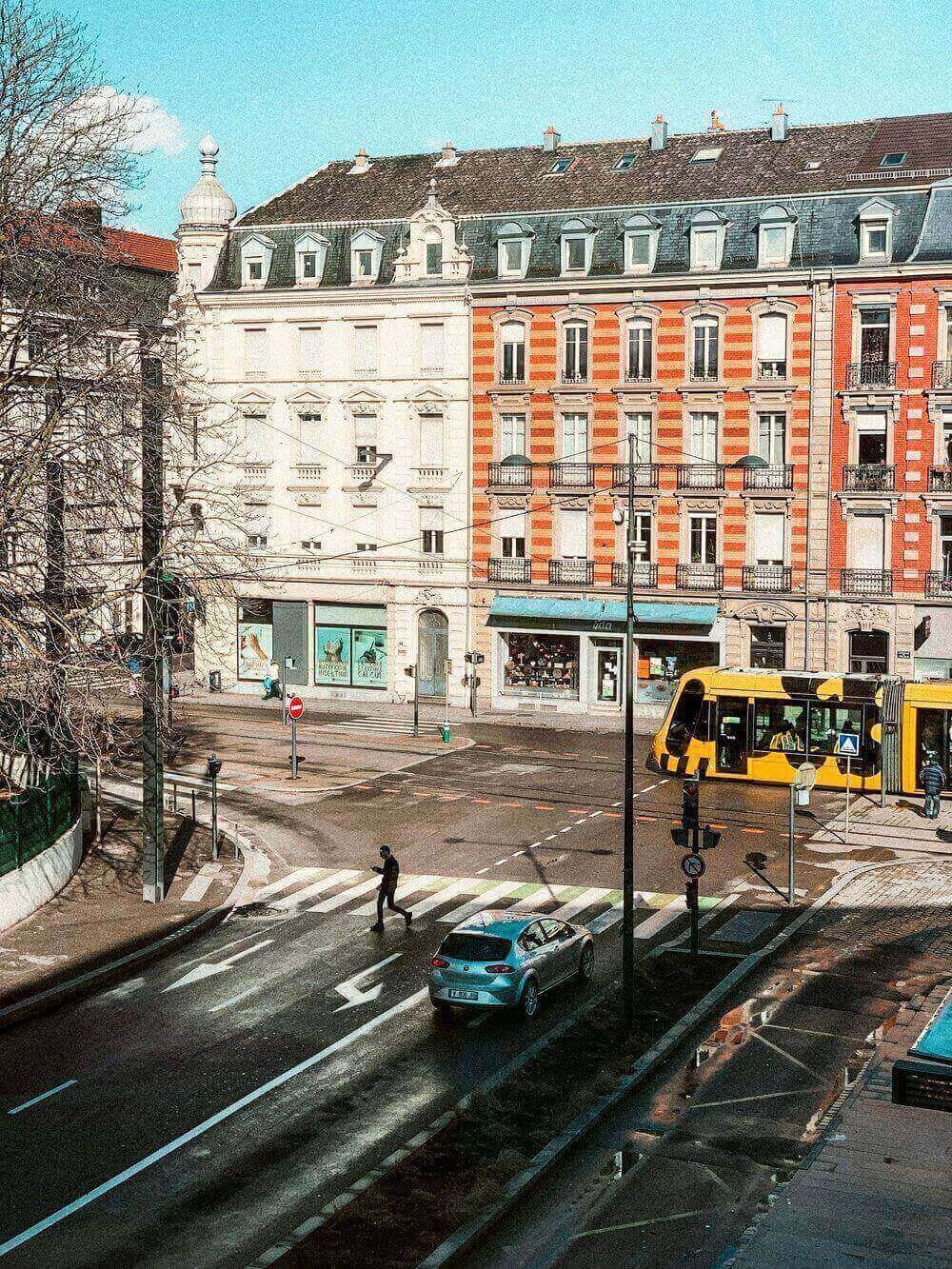 Vue panoramique d’une rue animée à Mulhouse, avec un tramway jaune et noir traversant l’intersection et des bâtiments aux façades colorées. Idéal pour découvrir comment devenir mannequin d’essayage dans une ville dynamique et cosmopolite.