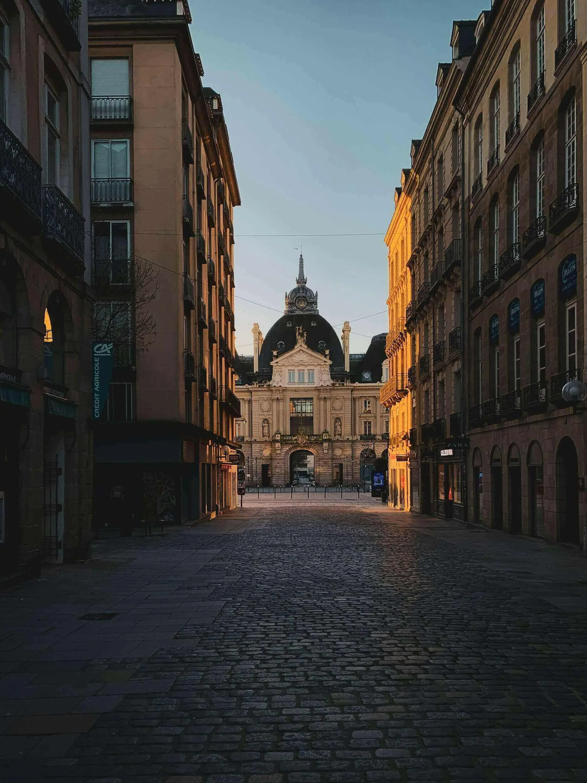 Vue d’une rue pittoresque à Rennes, bordée d’immeubles historiques et pavées de pierres, avec un édifice imposant au fond. Idéal pour découvrir notre agence et ses services d’essayage dans cette ville riche en architecture et culture.