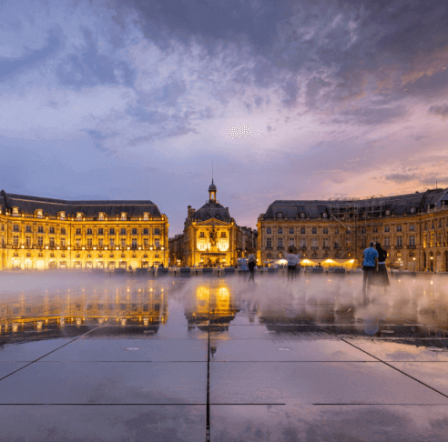 Vue de la Place de la Bourse à Bordeaux, avec un miroir d’eau reflétant les bâtiments historiques et le ciel crépusculaire. Idéal pour découvrir notre agence et ses services d’essayage dans cette ville élégante et culturelle.