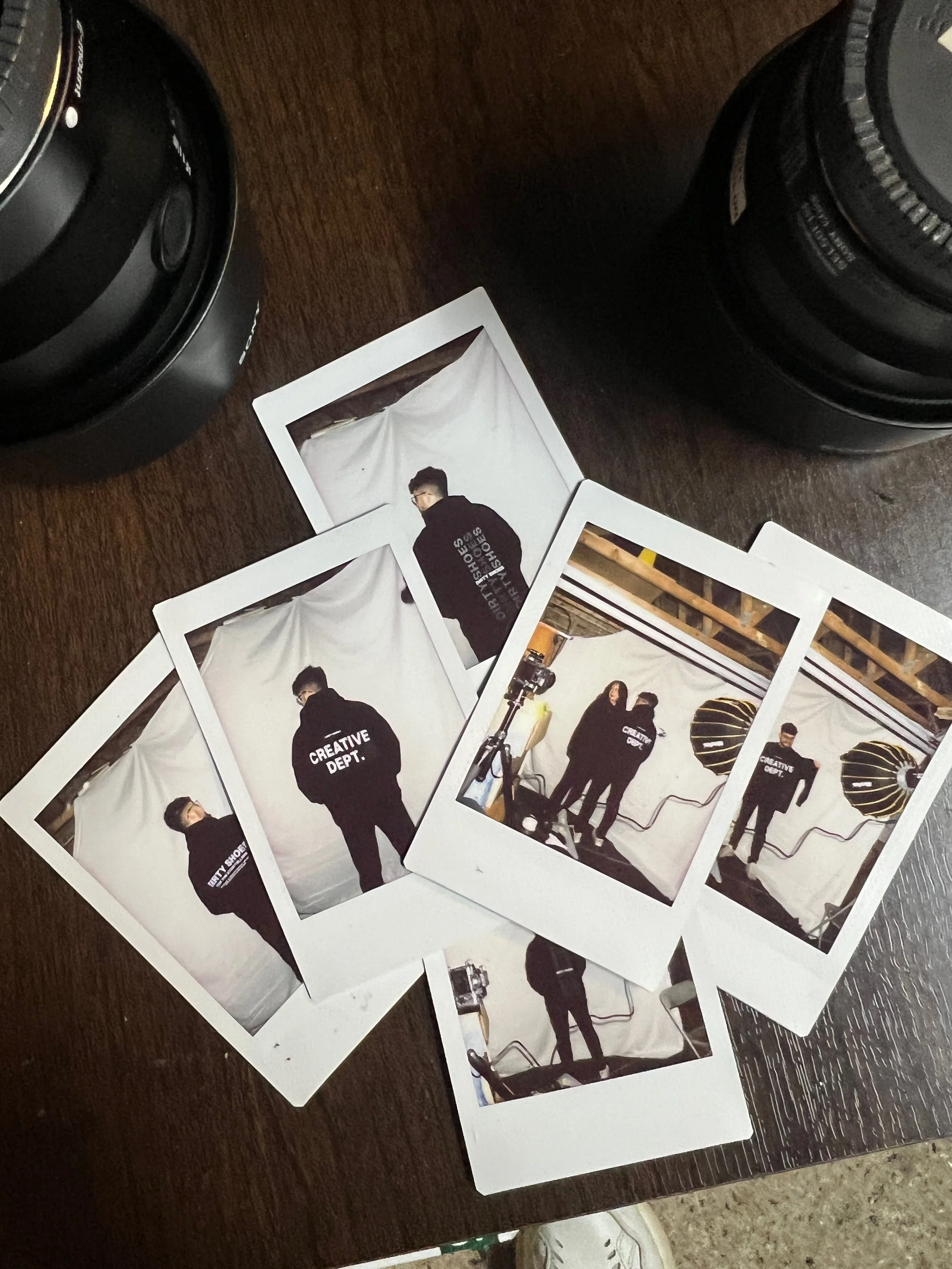 Polaroid photos on a wooden table with camera lenses, featuring people in "Creative Dept." hoodies, in a studio setting.