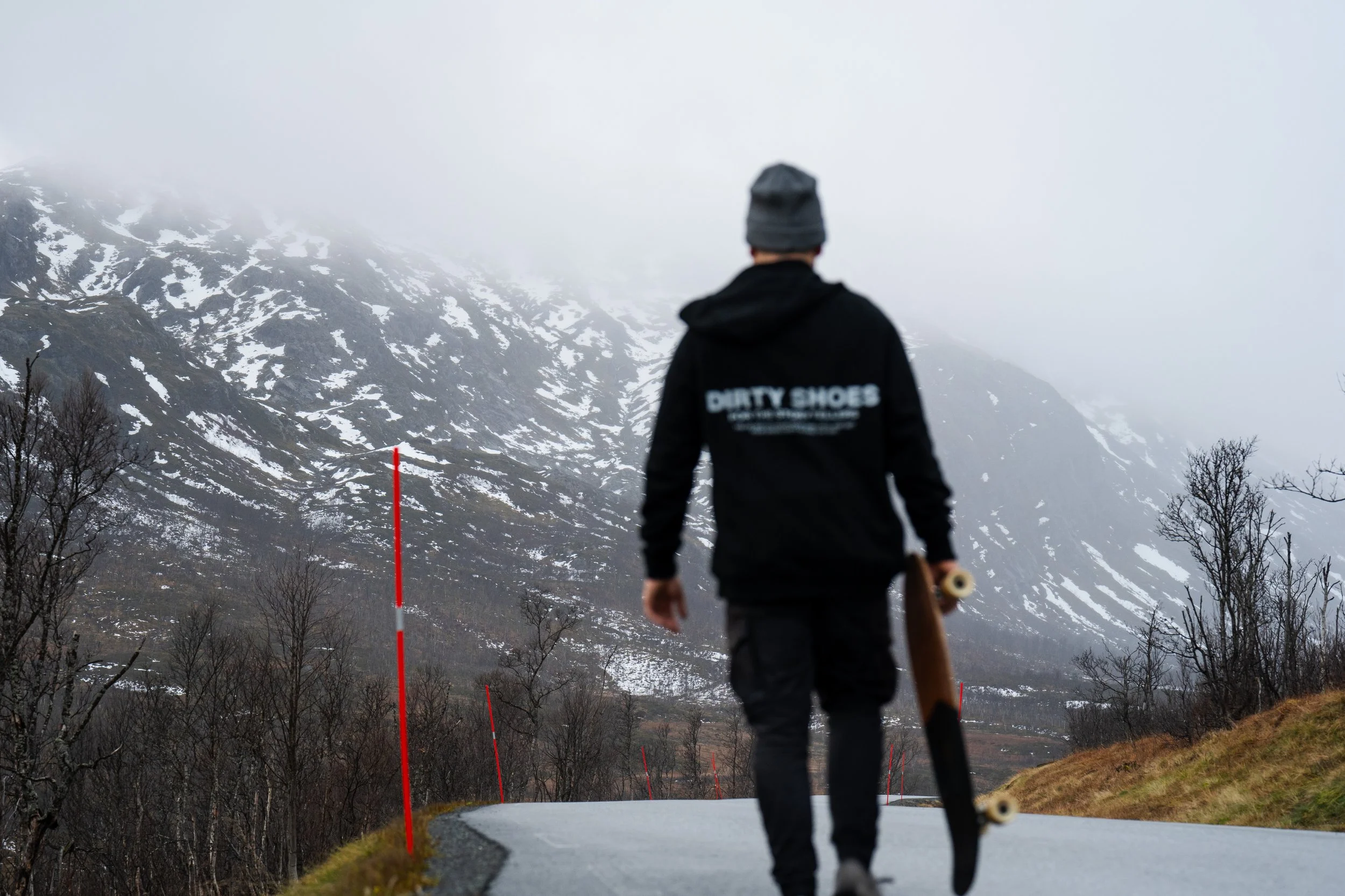 Person holding a skateboard walking on a foggy mountain road. Wearing a DIRTY SHOES hoodie.