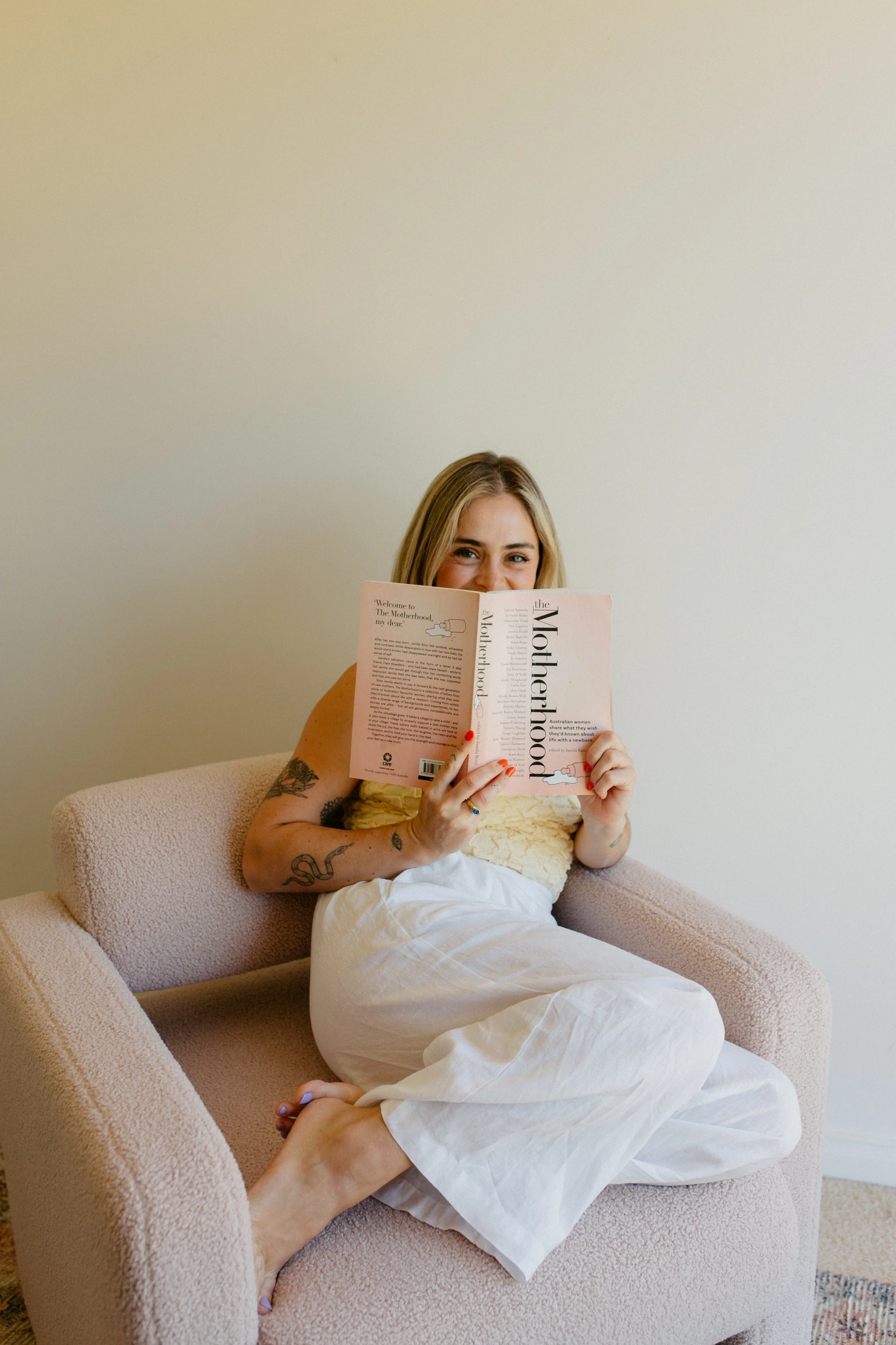 A woman with tattoos sitting on a pink armchair, smiling and reading a book titled "Motherhood" in a room with a plain white wall.