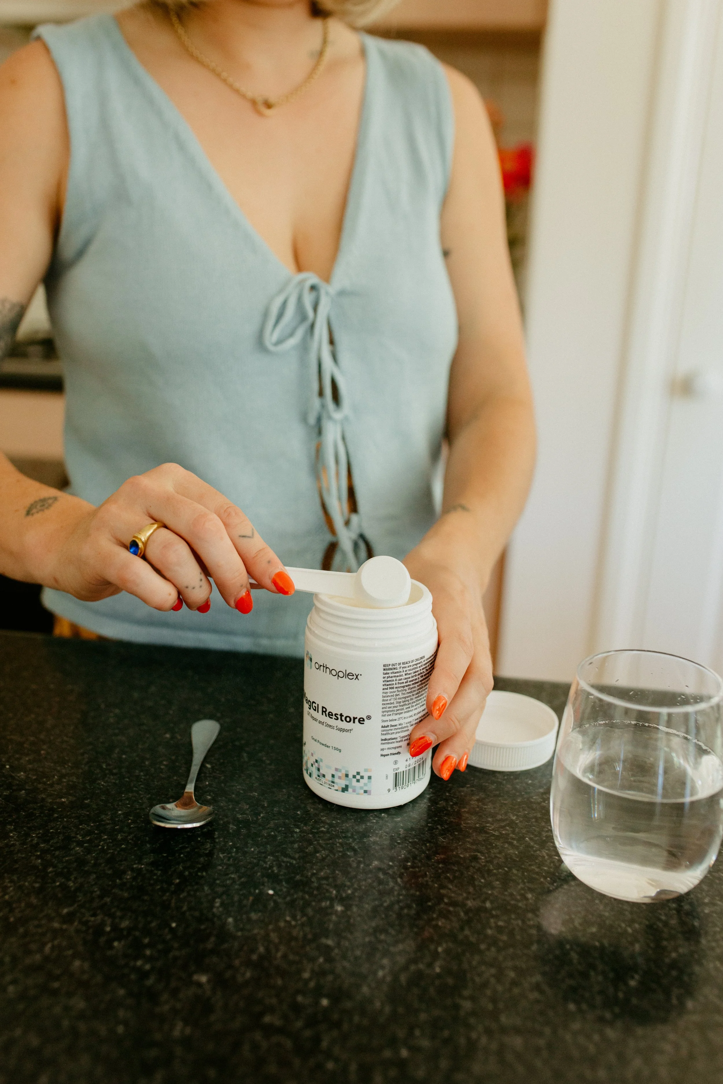 A woman with red nail polish opening a bottle of Orthoplex Veggi Restore supplement on a black kitchen countertop, with two glasses of water nearby.