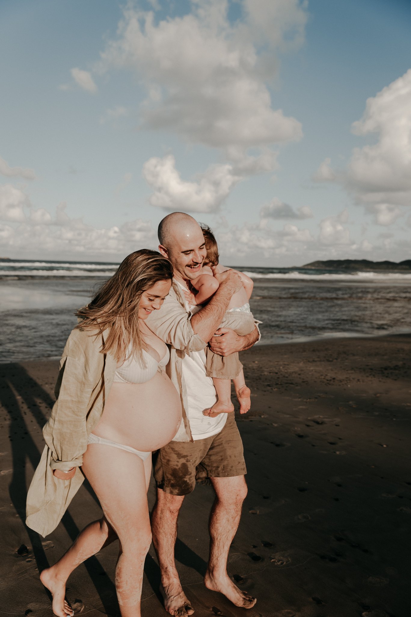 A family of three on a beach, smiling and holding a child, with a cloudy sky and ocean in the background.