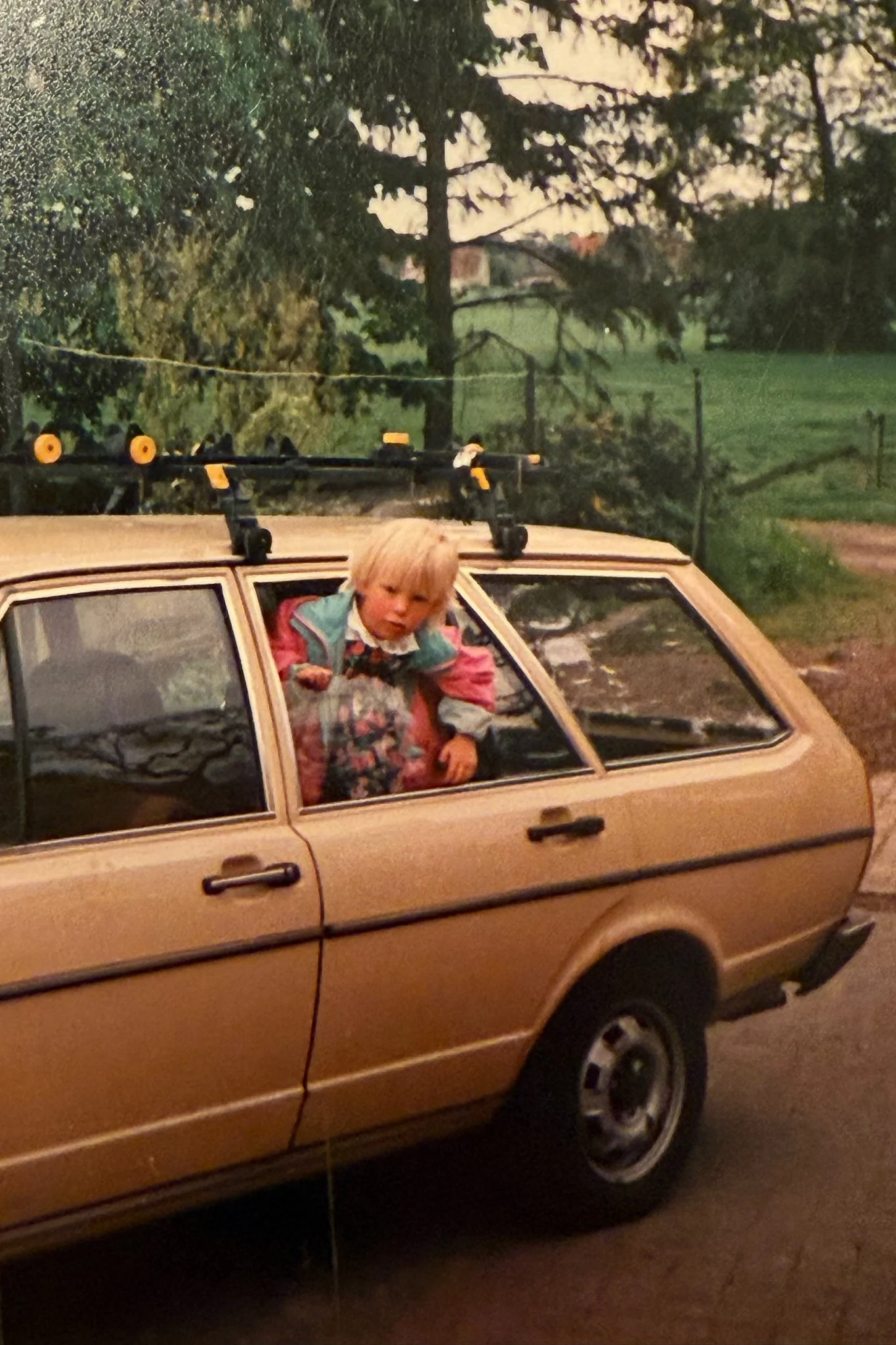 Ein Mädchen sitzt in einem braunen Auto und schaut neugierig aus dem Fenster, im Hintergrund sind Bäume und eine grüne Wiese zu sehen.