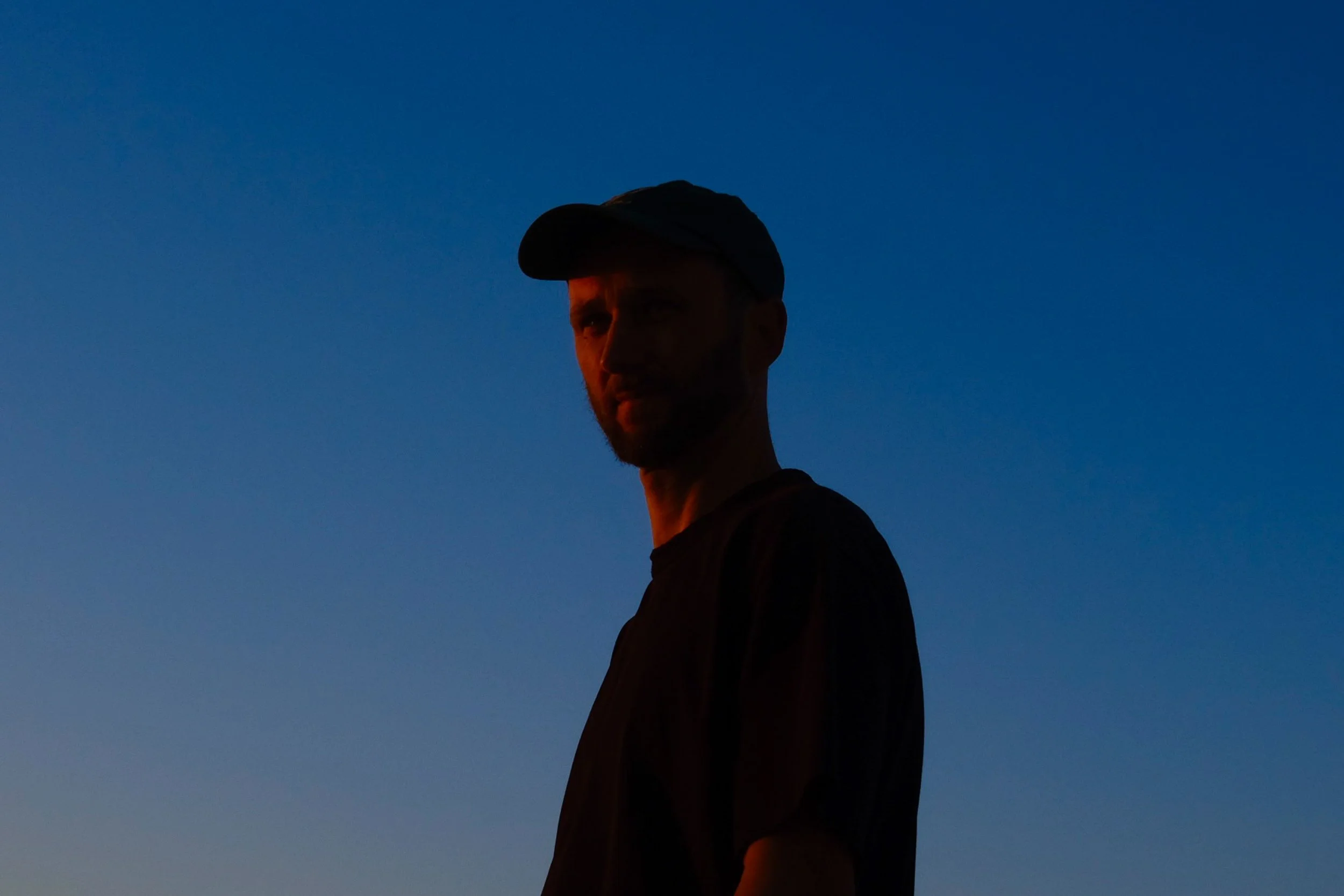 Porträt eines Mannes mit dunklen Haaren, Bart und Mütze, der im Sonnenuntergang schaut, gegen einen blauen Himmel.