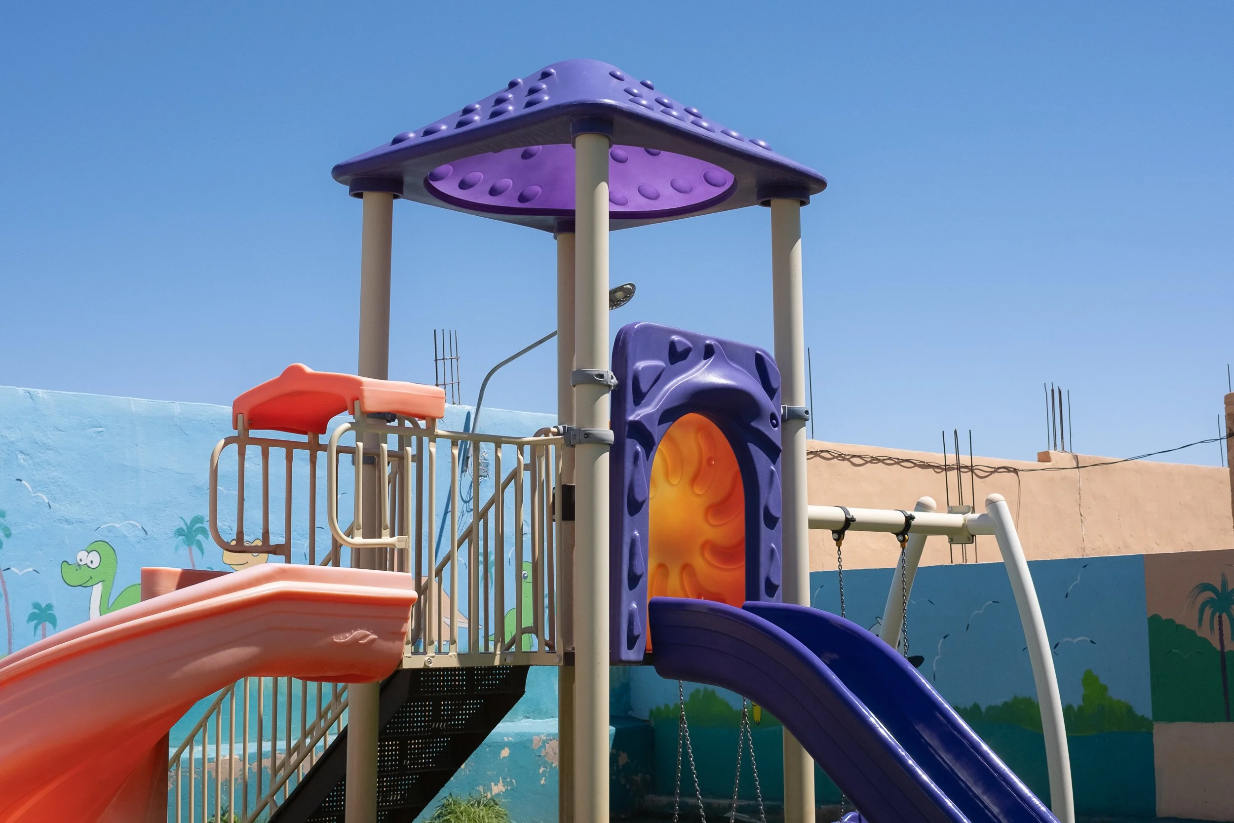 Bunte Kinderspielplatz mit Rutschen, Kletterturm und Schaukeln, vor bunter Mauer mit Tiermalerei, bei blauem Himmel.