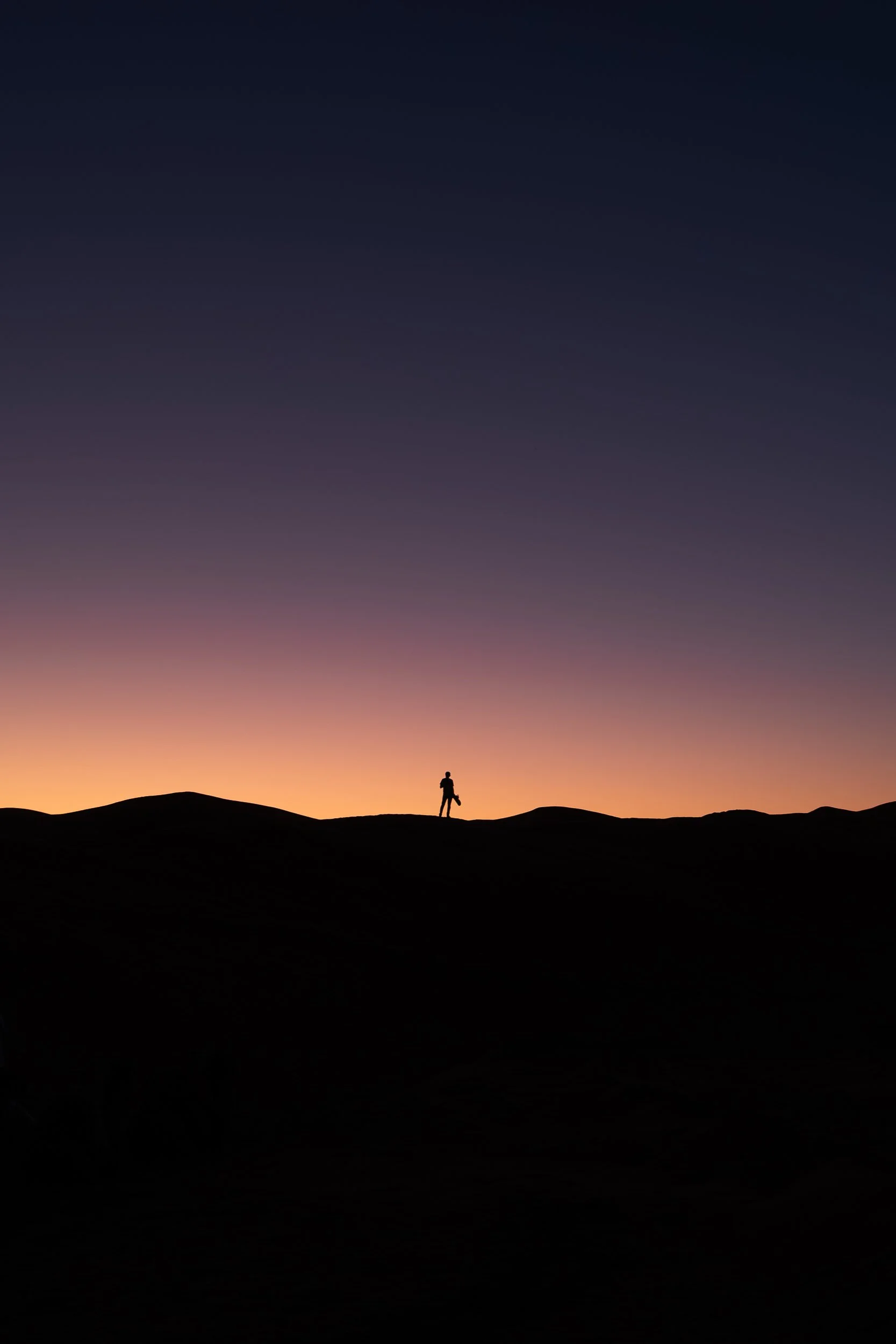 Ein einzelner Mensch steht auf einem Hügel in einer Wüstenlandschaft bei Sonnenuntergang, silhouettiert gegen den mehrfarbigen Himmel mit einem Farbverlauf von Orange bis Dunkelblau.