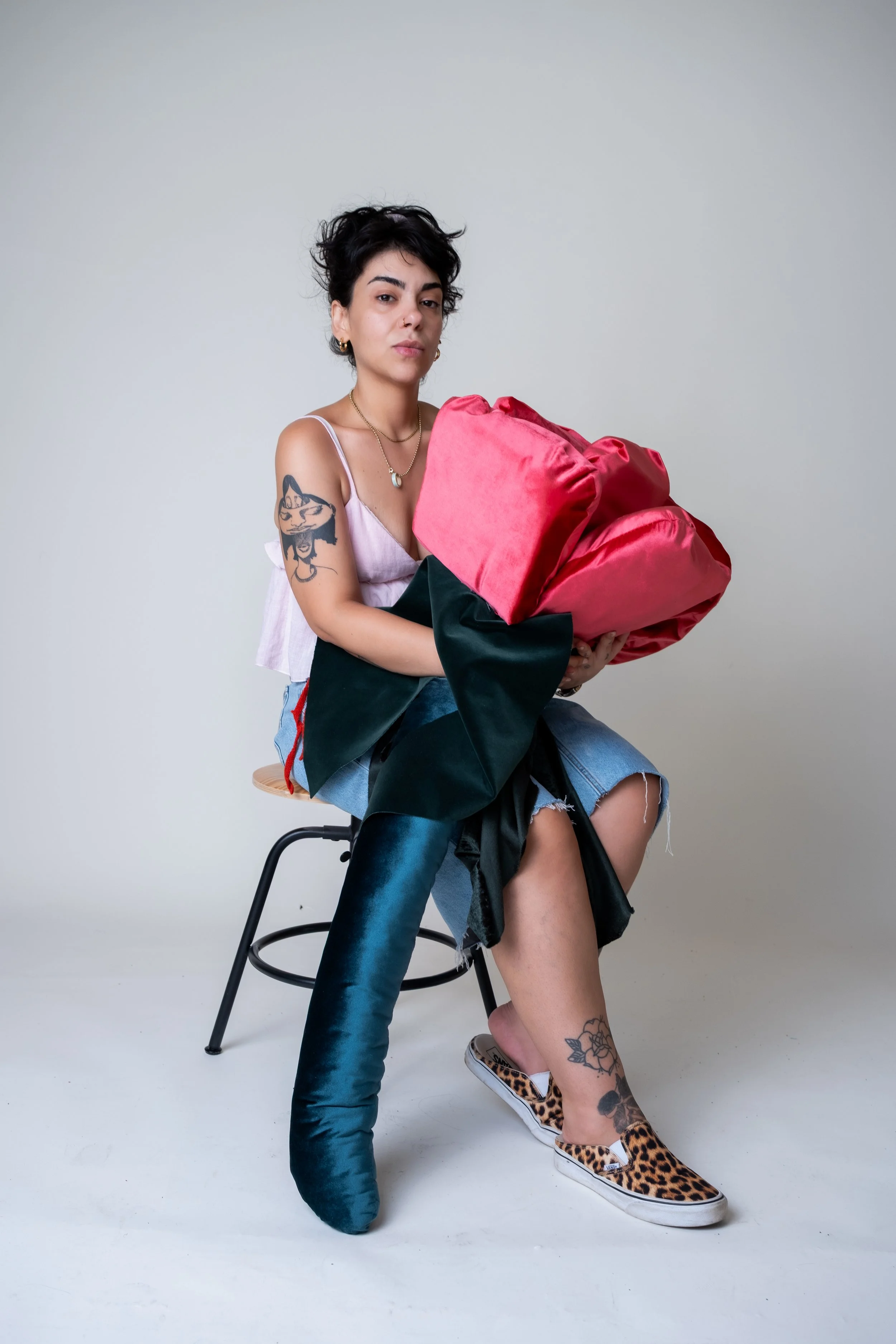 Frau sitzt auf einem Hocker, trägt Jeans, einen weißen Tank-Top, auffällige Schuhe mit Leopardenmuster, hat Tattoos am Arm und Bein, hält eine große pinkfarbene Tasche, hat kurze schwarze, lockige Haare, trägt kleine Goldohrringe und eine Halskette, 