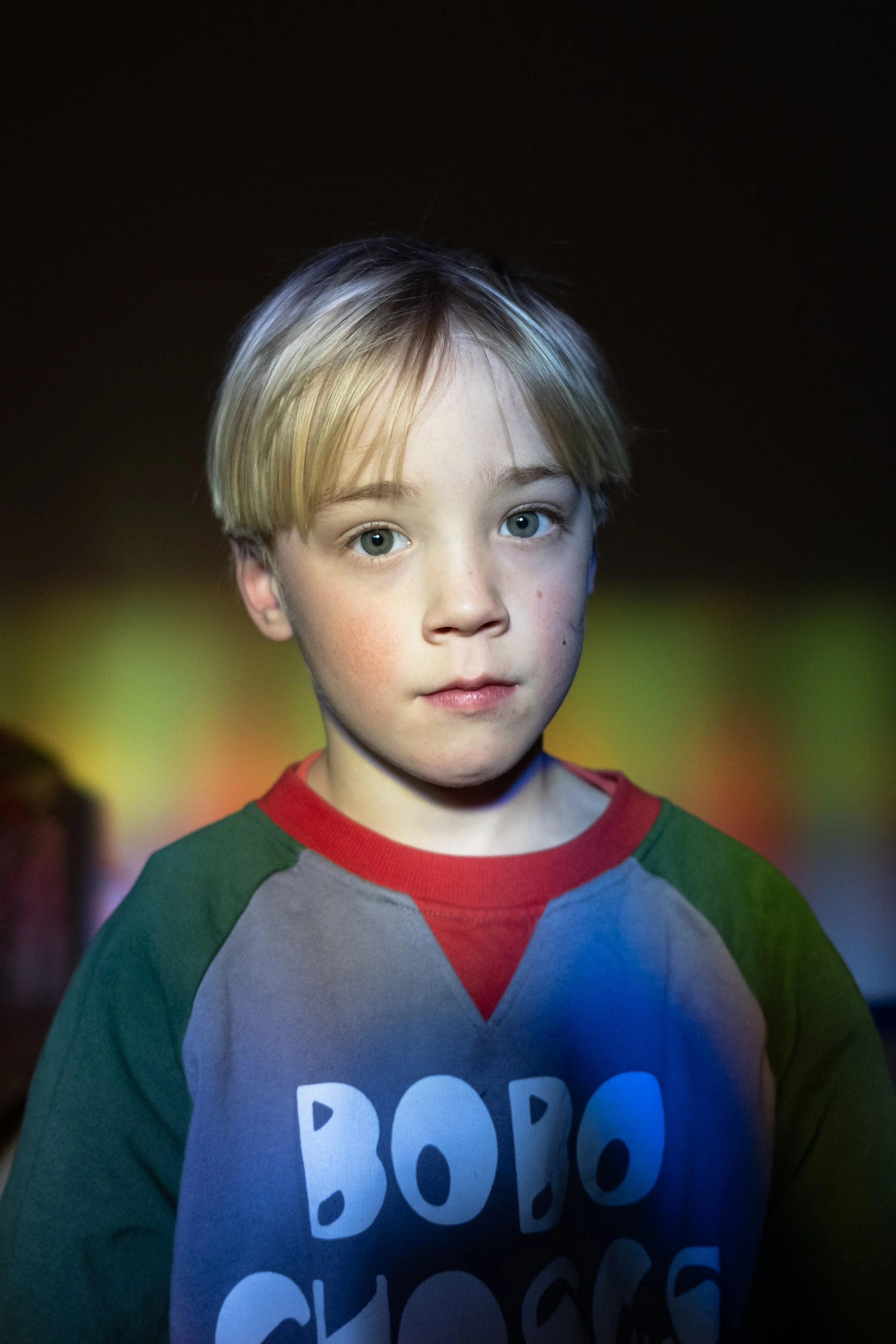 Ein Junge mit blonden Haaren und blauen Augen trägt ein graues Sweatshirt mit grünen Ärmeln und einer roten Halskante. Der Hintergrund ist dunkel, und es gibt einen Farbverlauf aus warmen und kühlen Tönen.