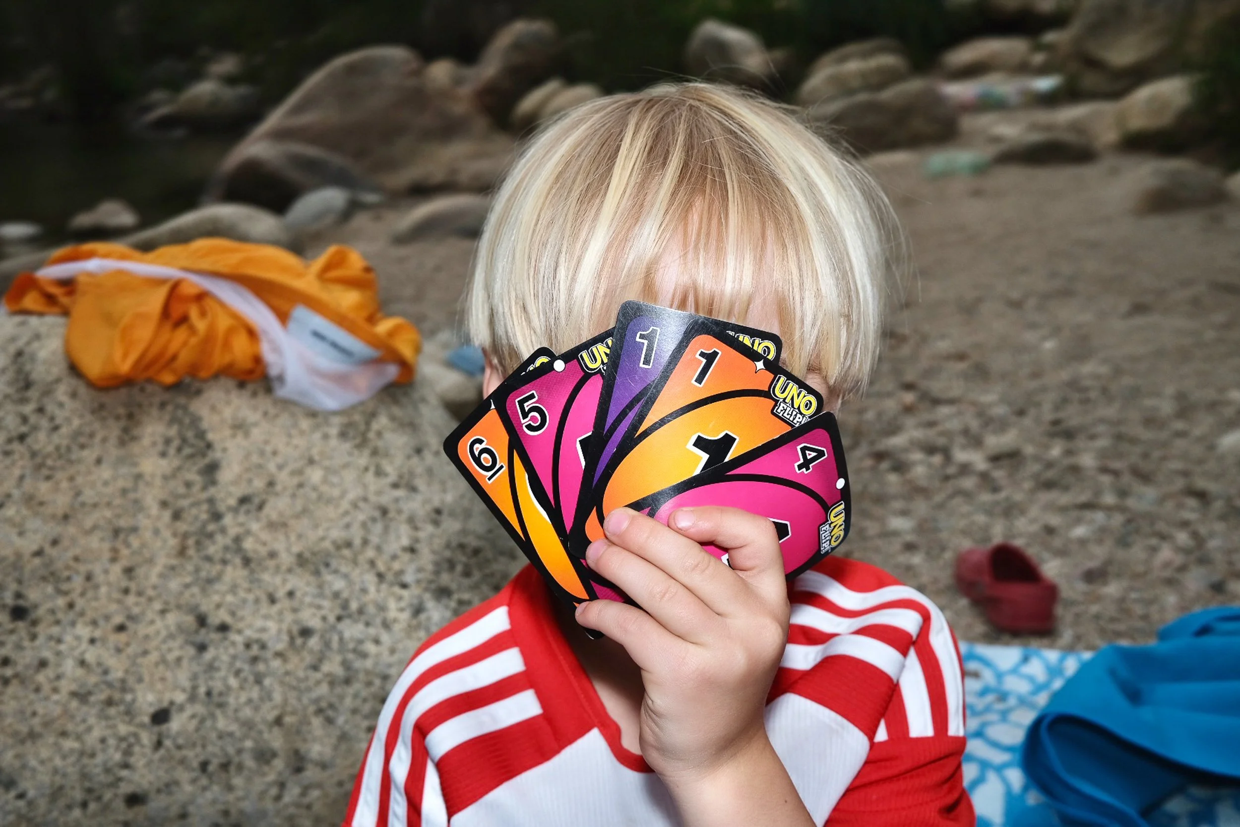 Ein Kind mit blonden Haaren, das bunte UNO-Karten hält, sitzt an einem Strand mit Steinen und Wasser im Hintergrund, trägt ein rotes und weiß gestreiftes Hemd.