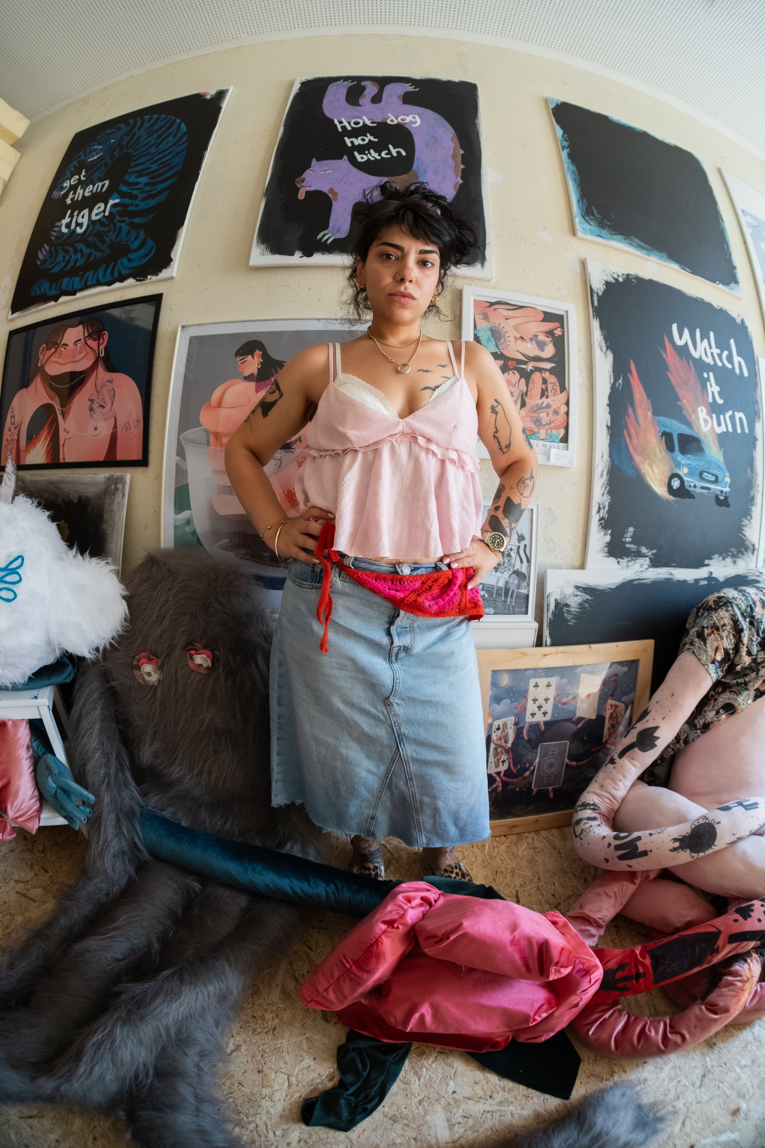 Eine junge Frau mit dunklen, zerzausten Haaren steht vor einer Wand mit verschiedenen Kunstwerken, darunter ein Bild mit einer Schlange und der Text „Get them tiger“, ein Bild eines Dinosauriers mit dem Text „Hot dog hot bitch“ und ein Bild mit brenn