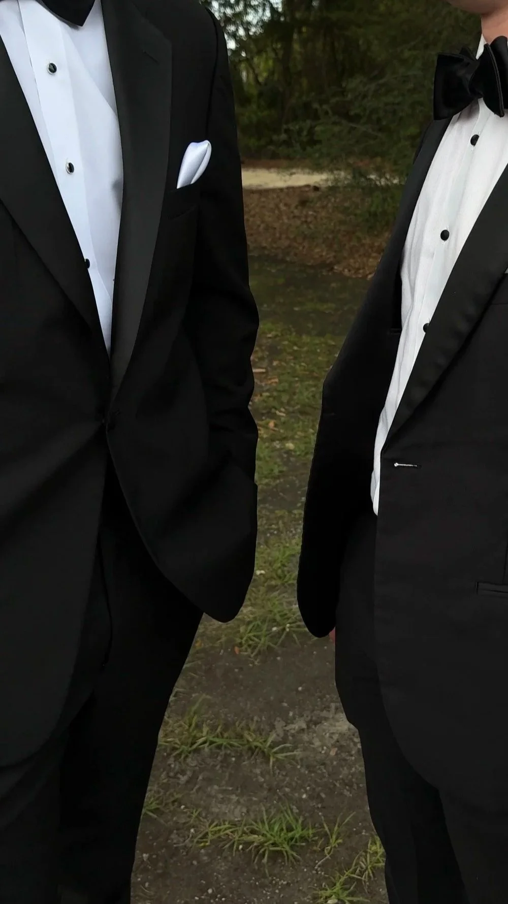 Close-up of two men in black tuxedos and white dress shirts, facing each other outdoors, with blurred trees and plants in the background.