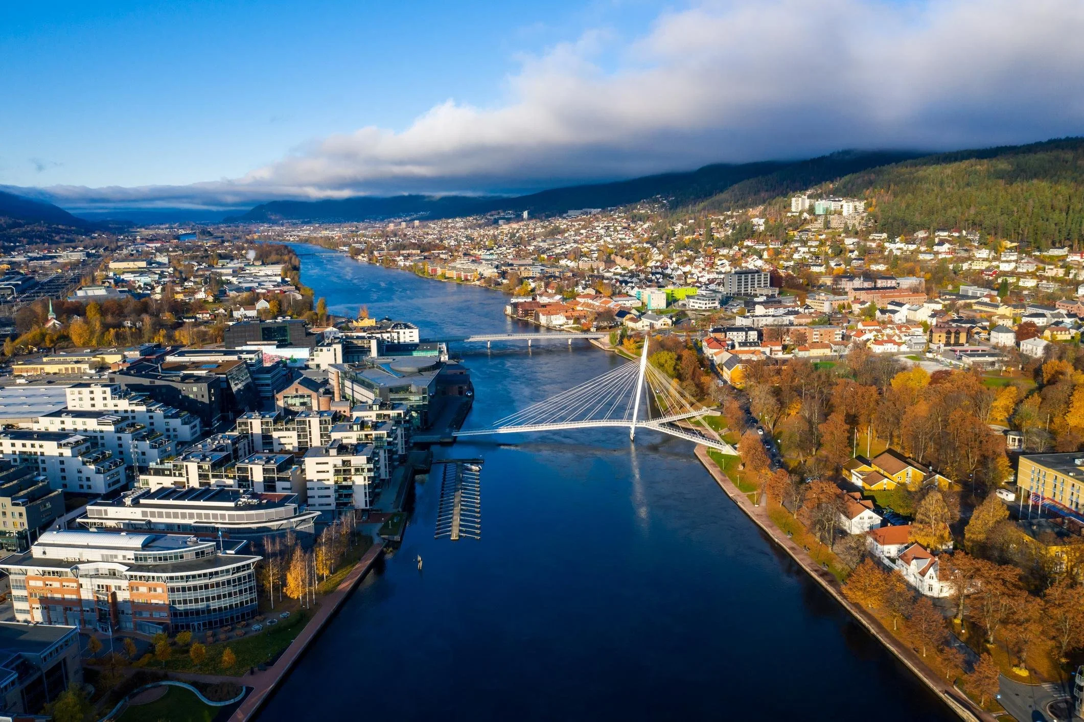 Aerial view of a city with a river running through it, surrounded by hills and mountains, with a bridge crossing the river and buildings on both sides, under a sky with clouds.