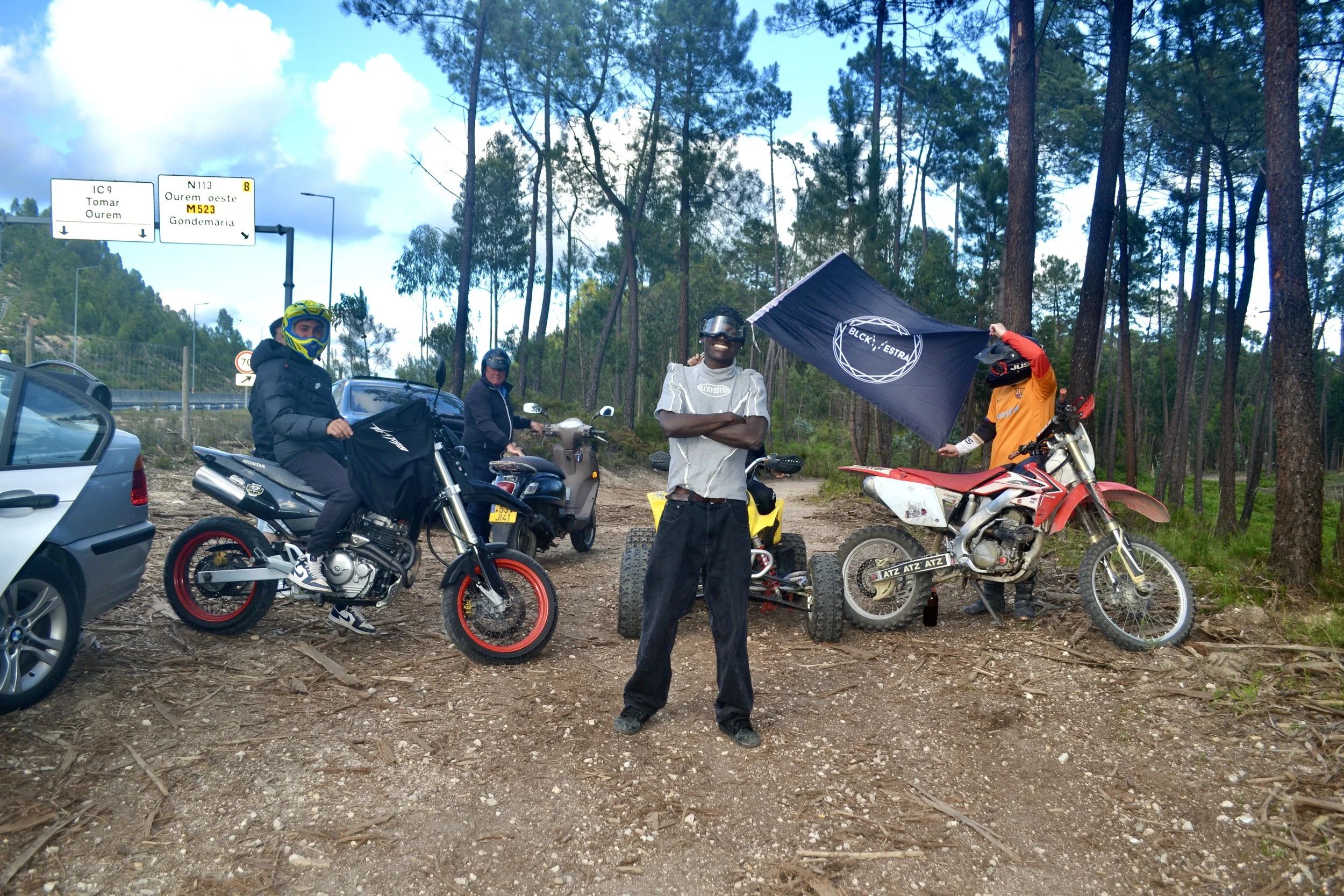 Four men are in a forested area with two motorcycles and a quad bike. One man stands in the center with arms crossed, smiling at the camera. The other three men are standing with one holding a flag. Cars and a scooter are parked behind them. Signage 