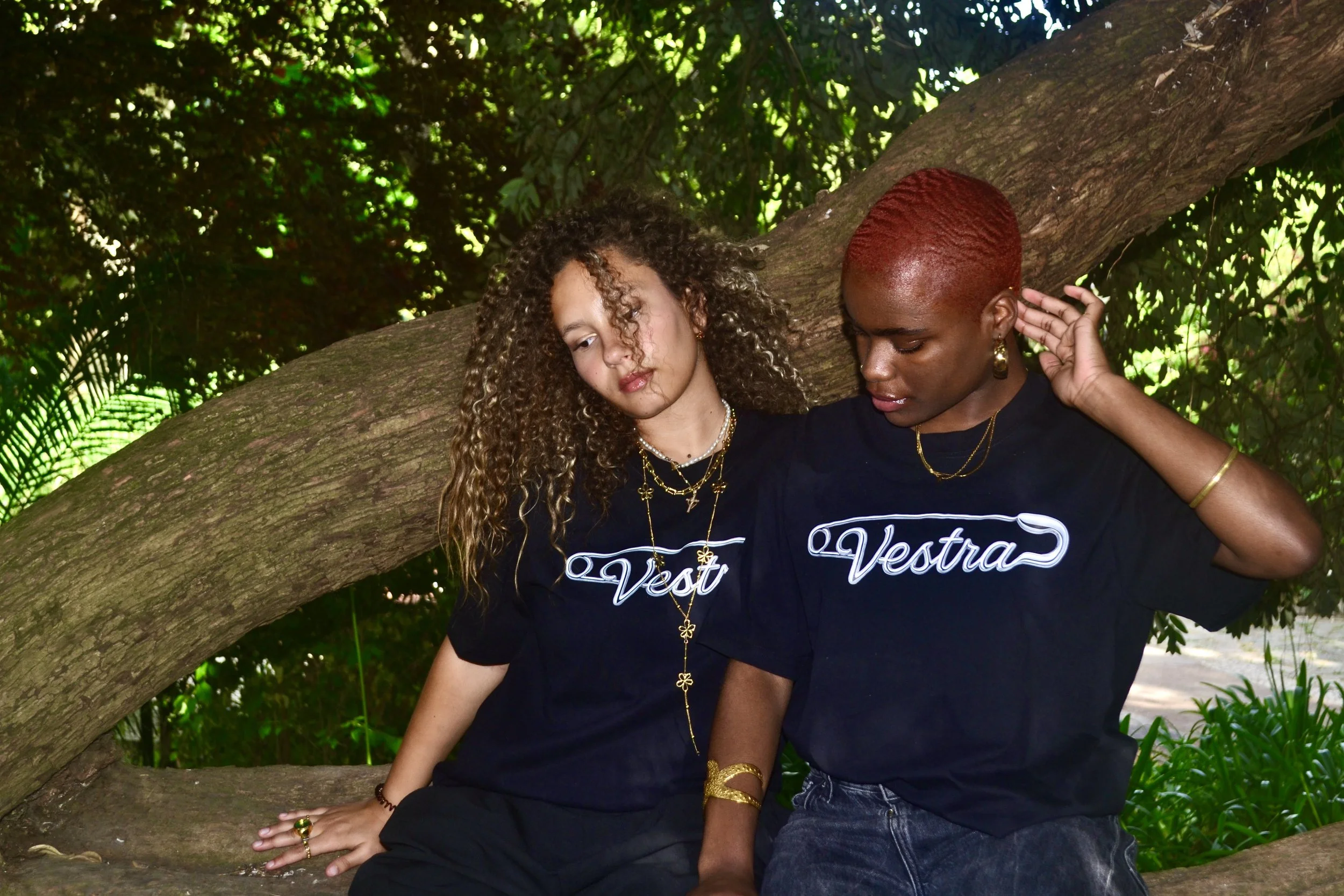 Two women sitting on a branch of a large tree, both wearing black shirts with the word "Vestra" on them, surrounded by green foliage.