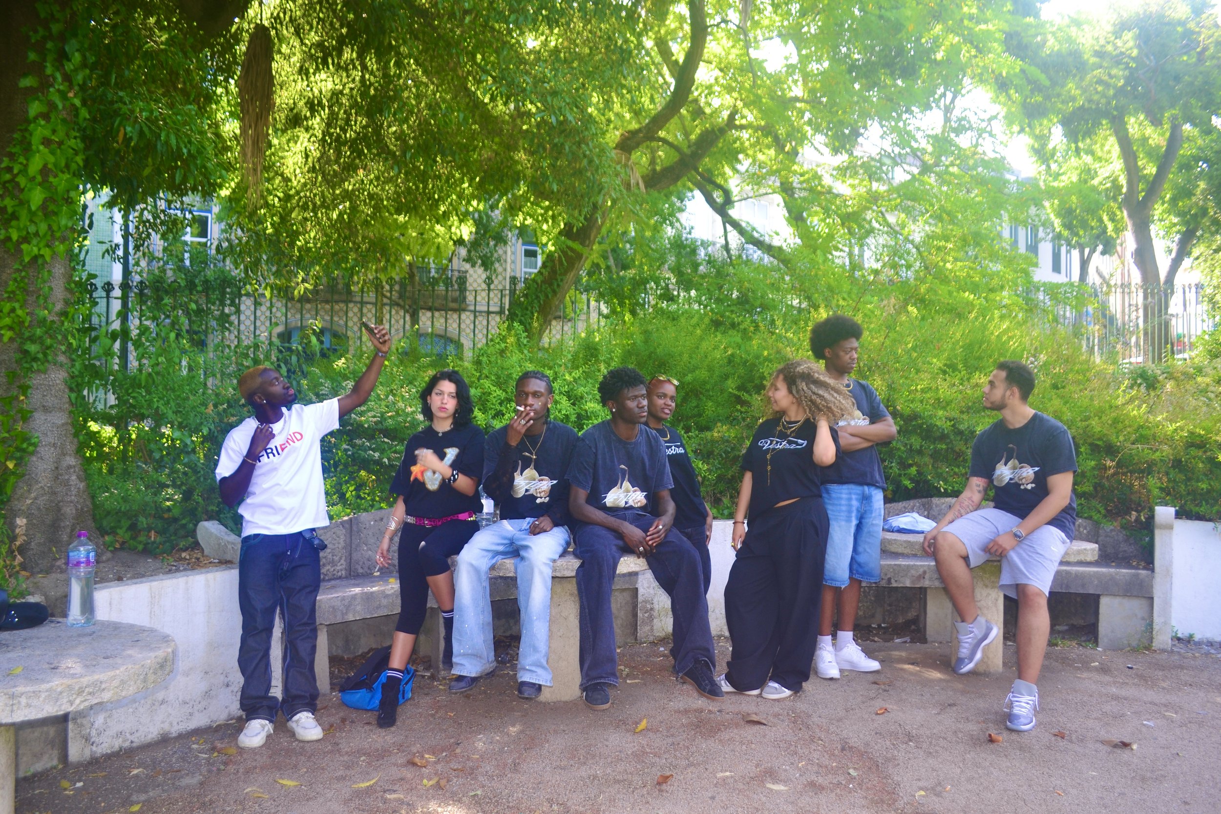 Group of people sitting and standing on a park bench surrounded by green trees and foliage, with some holding drinks and taking photos.