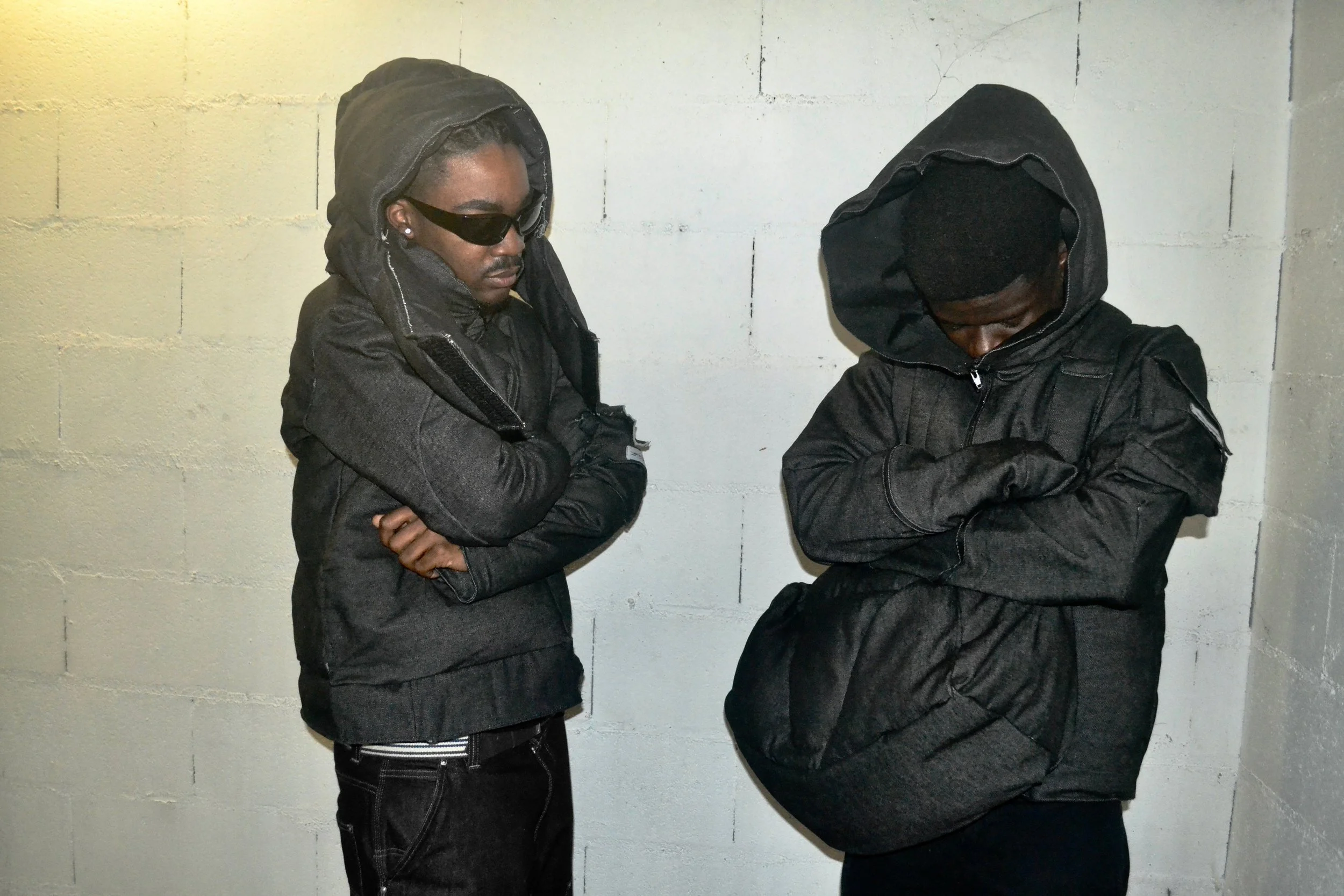 Two young men dressed in black hooded jackets with arms crossed, standing against a white brick wall, appearing to be in a contemplative or serious mood.