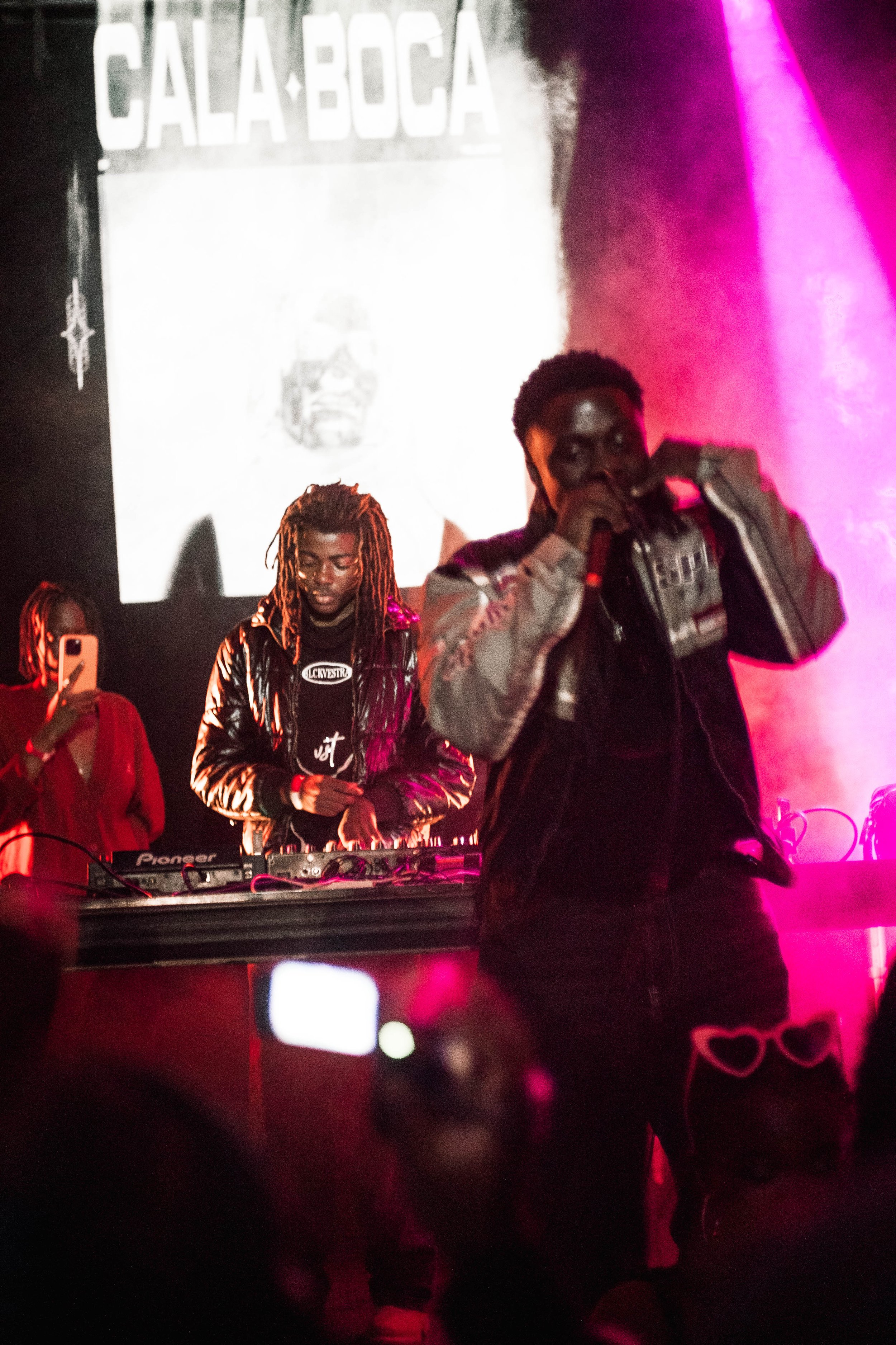 A live performance at Calaboca nightclub with two male performers in the foreground, one speaking into a microphone and the other working DJ equipment, under pink and purple stage lighting, with a woman on the left taking a photo.