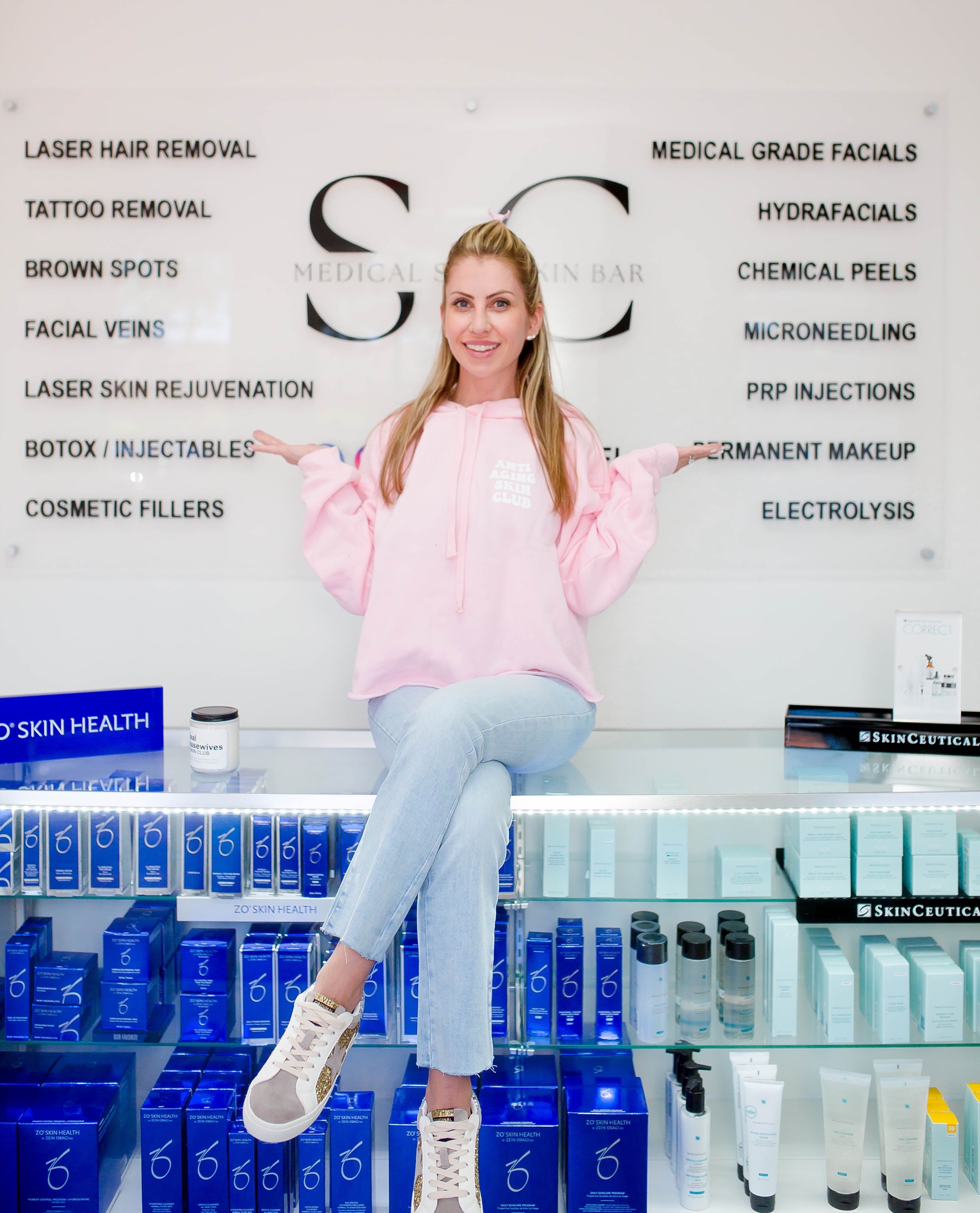 Woman in a pink hoodie sitting on a shelf displaying skincare products with a sign for various skin treatments in the background.