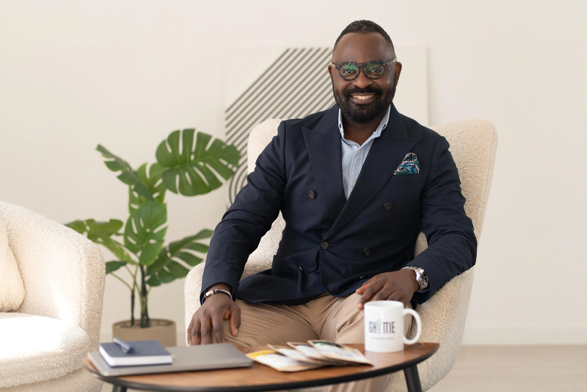A man sitting in a beige armchair, wearing glasses, a navy blue blazer, a light blue shirt, and tan pants. He is smiling, with a mug, notebooks, and brochures on a wooden table in front of him. There is a green plant and abstract black and white artwork in the background.