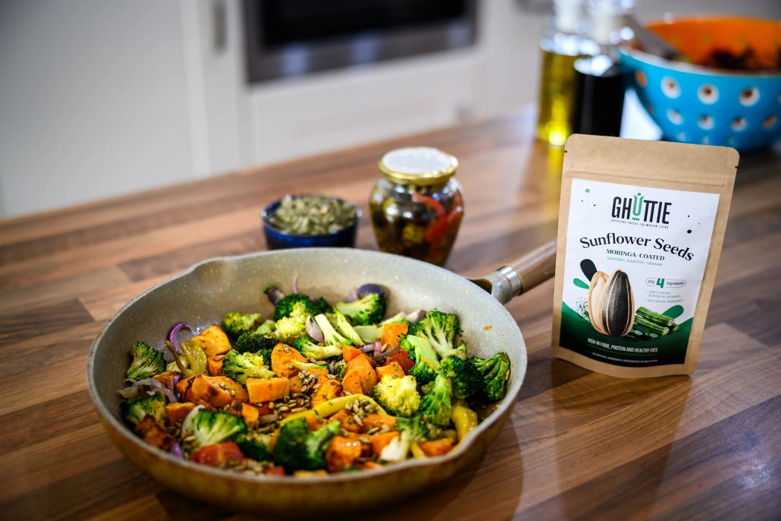 A frying pan filled with cooked broccoli, sweet potatoes, and onions on a wooden kitchen counter. Behind the pan, there are two jars and a package of "Ghutti Snacks" sunflower seeds.