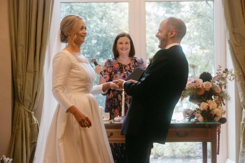 Bride in white dress and groom in a tux exchange rings, with celebrant in pink flowery dress and microphone between who's smiling and groom is leaning back and laughing.