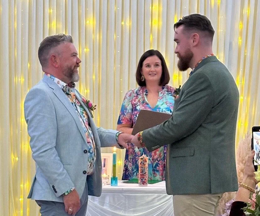 Two men, one in a blue jack and one in a green jacket exchange vows & rings, with the celebrant in a flowery dress between them.