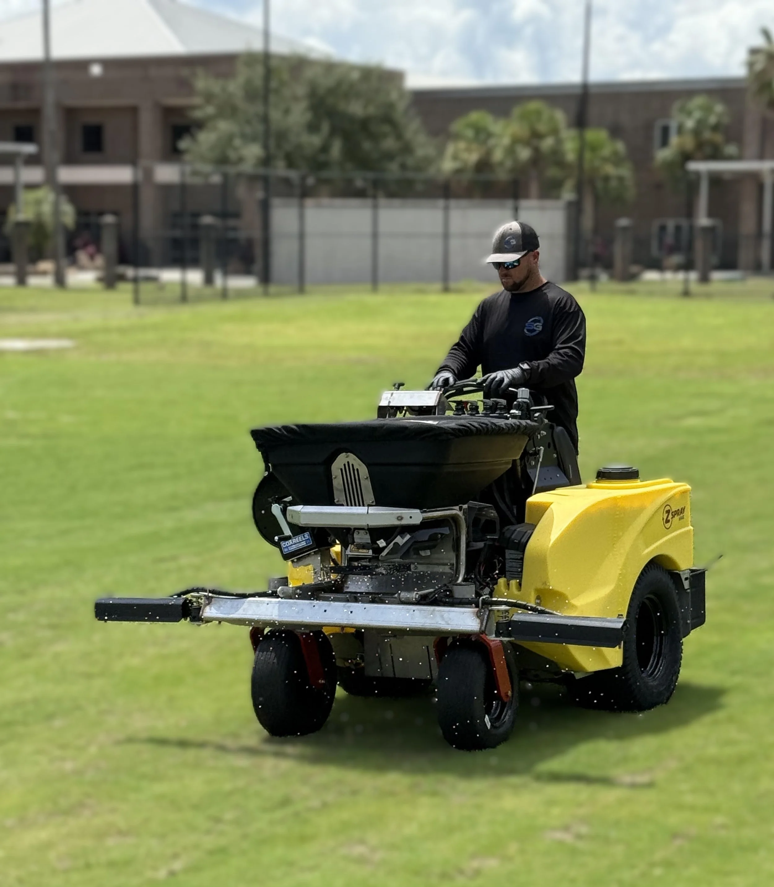A man operating a yellow and black lawn mower on a grassy area, wearing sunglasses, a black long-sleeved shirt, a cap, and gloves, with a fence and residential buildings in the background.