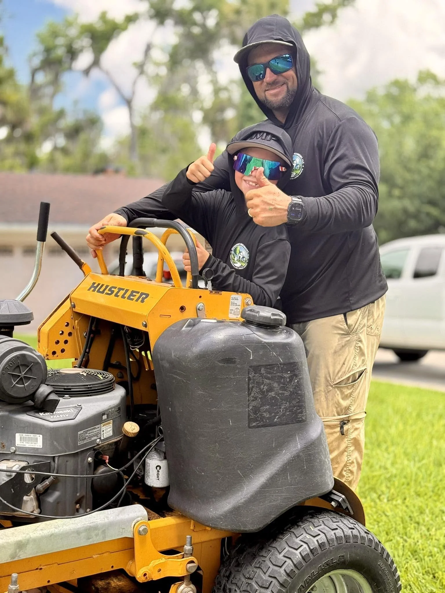 Two men in black jackets with logos, one wearing sunglasses and a cap, and the other also with a cap and reflective sunglasses, posing with a yellow Hustler ride-on lawnmower outdoors.