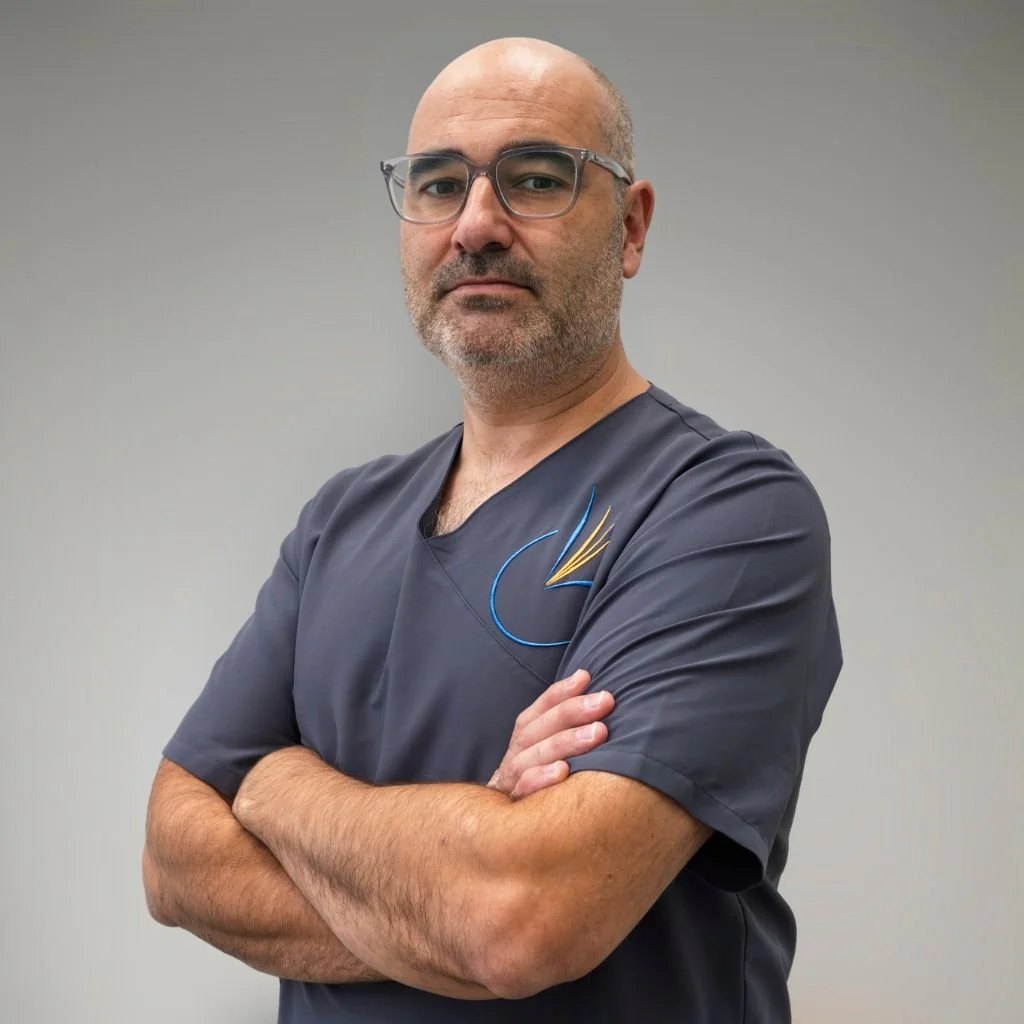 Hombre con gafas y barba, vestido con uniforme de trabajo, de postura confiada y con brazos cruzados, frente a un fondo gris.