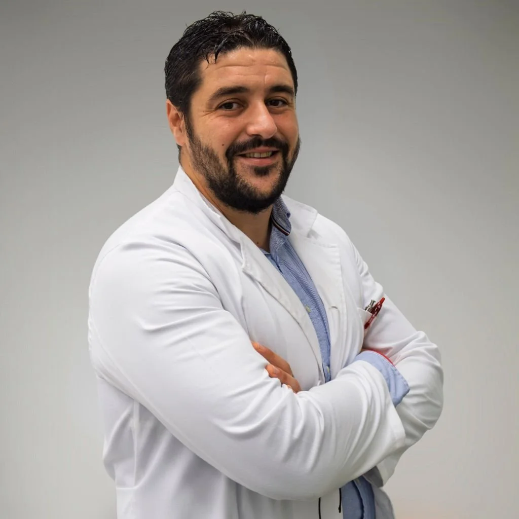Retrato de un hombre con barba y bigote, vestido con bata blanca, sonriendo y con los brazos cruzados, en un fondo de pared gris.