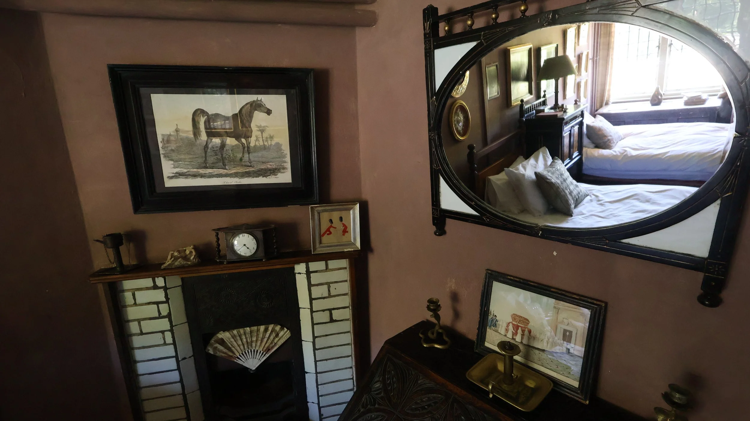 Interior of a vintage-style bedroom with a mirror reflecting a bed with pillows, framed artwork, a clock, and decorative items on shelves and tables.