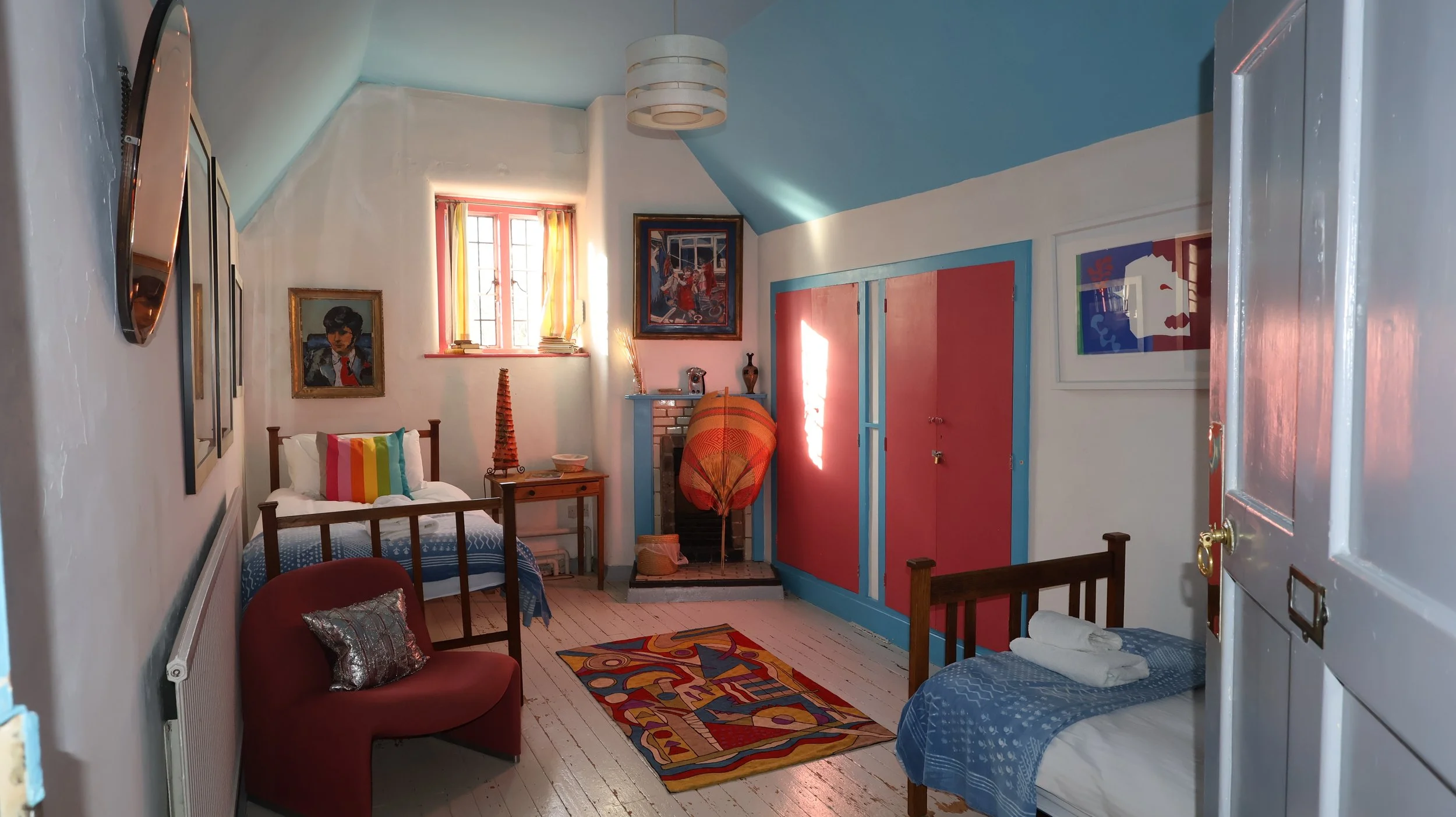 A colorful bedroom with two beds, one with rainbow striped pillow, and another with rolled towels. Bright sunlights through a window with pink curtains, a small wooden nightstand, a red and blue wardrobe, artwork on white walls, a painted rug, and de