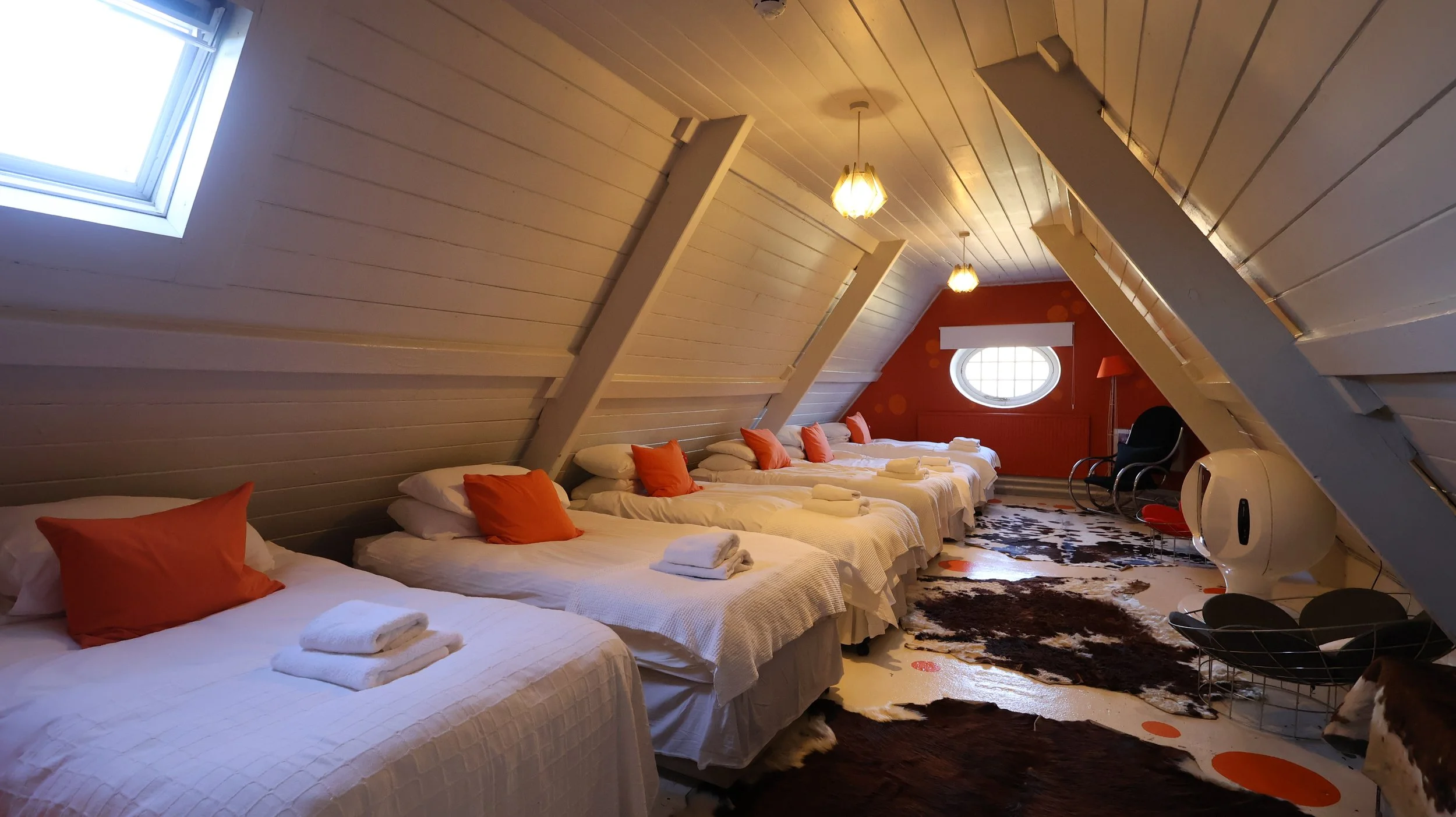 A cozy attic bedroom with six beds, each with white linens and orange pillows, under a sloped ceiling with skylight windows. There is a circular window at the far end, a black armchair with a red lamp, and a cowhide rug on the floor. country house 