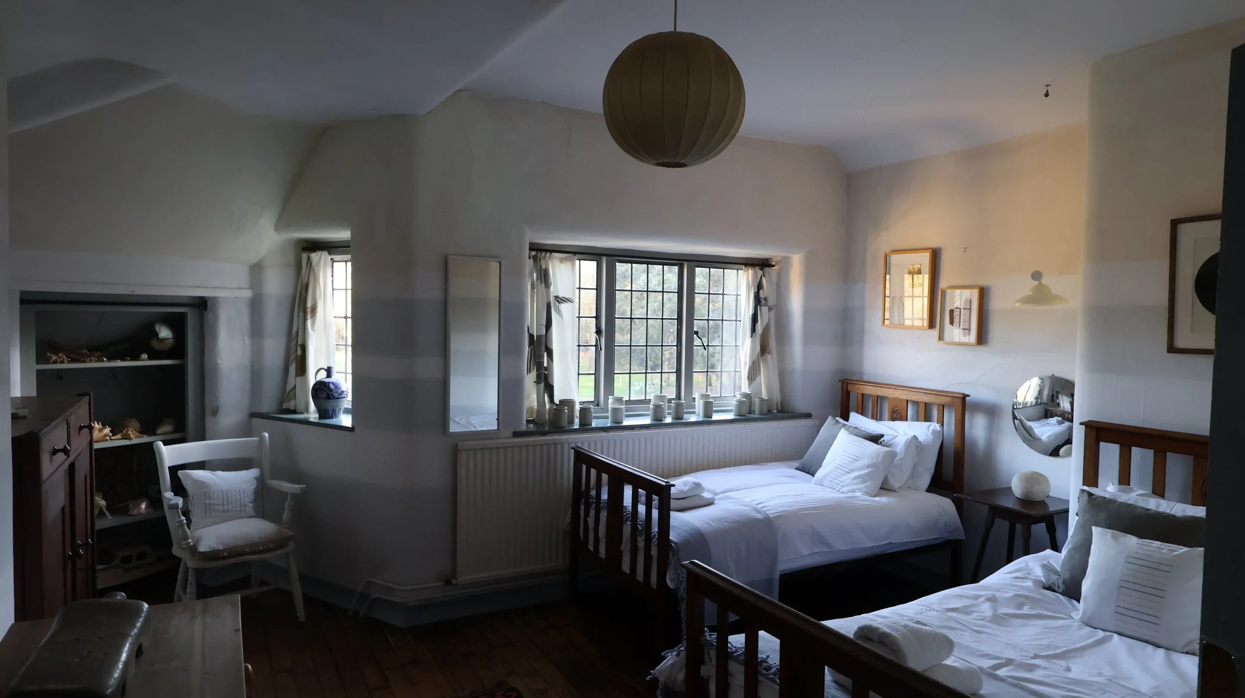 A cozy bedroom with two white beds, wooden headboards, and multiple pillows. A large window with a grid pattern lets in natural light, flanked by curtains. Decor includes framed pictures, a round mirror, a small table with a decorative object, and a 