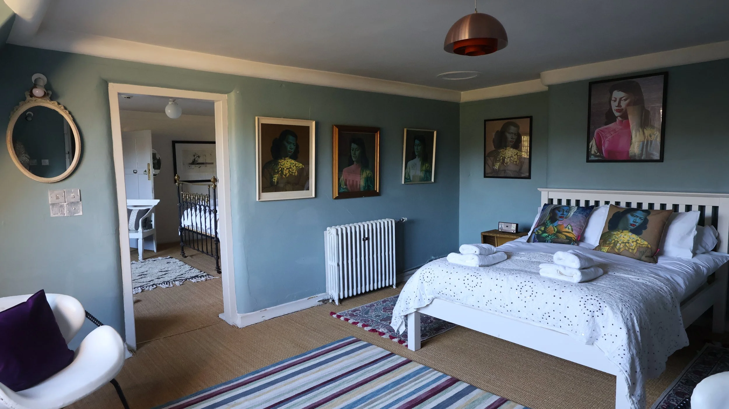 A bedroom with light blue walls featuring framed portraits, a white bed with embroidered linens and decorative pillows, and a small wooden side table with a clock. Towels are folded on the bed, and a striped rug is on the floor. There is a mirror and