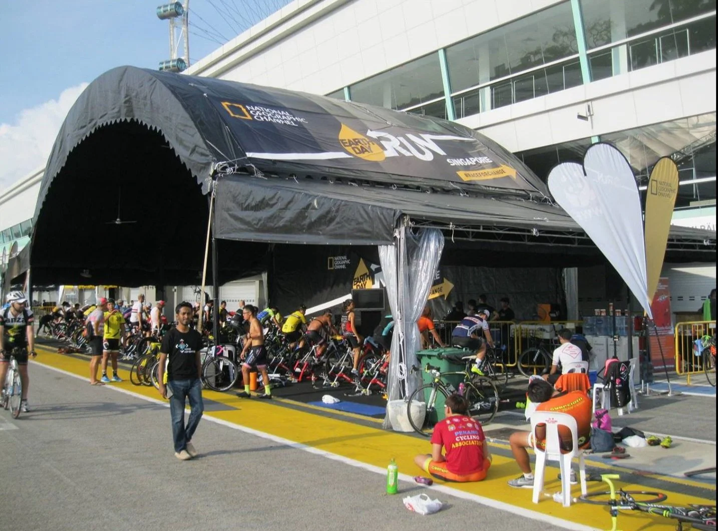 Cyclists preparing at a pit stop under a black tent during a race event, with bicycles and racing gear visible, at a modern sports facility.