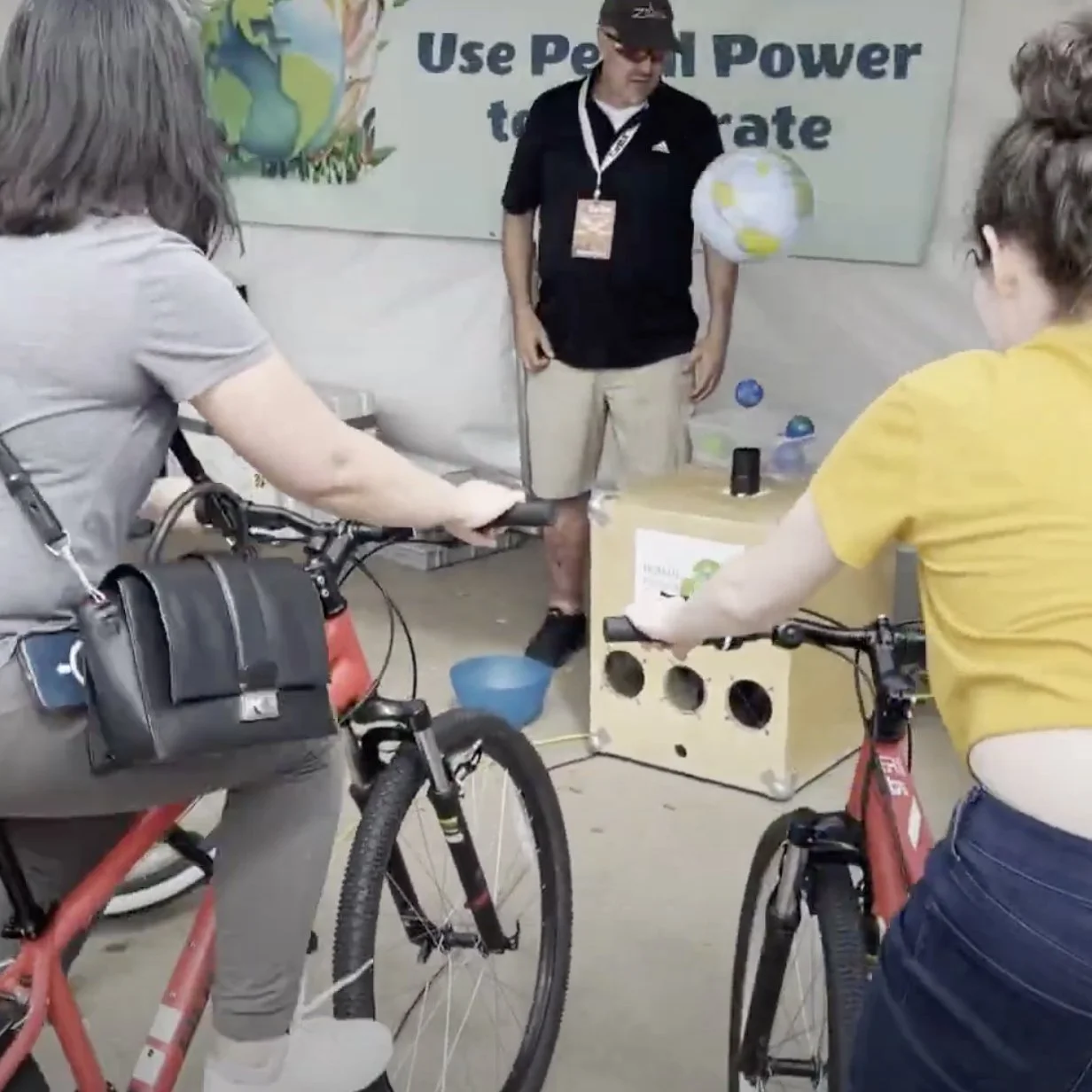 Young boy riding a yellow stationary bicycle at an outdoor event with several onlookers, including children and adults, nearby. A vendor setup with equipment and a man with a camera in the background.