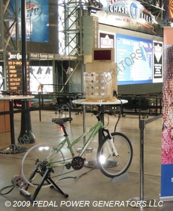 Indoor sports arena with a green bicycle mounted on a stationary trainer, positioned on a circular platform in front of a Bettenhausen score display and a large scoreboard in the background.