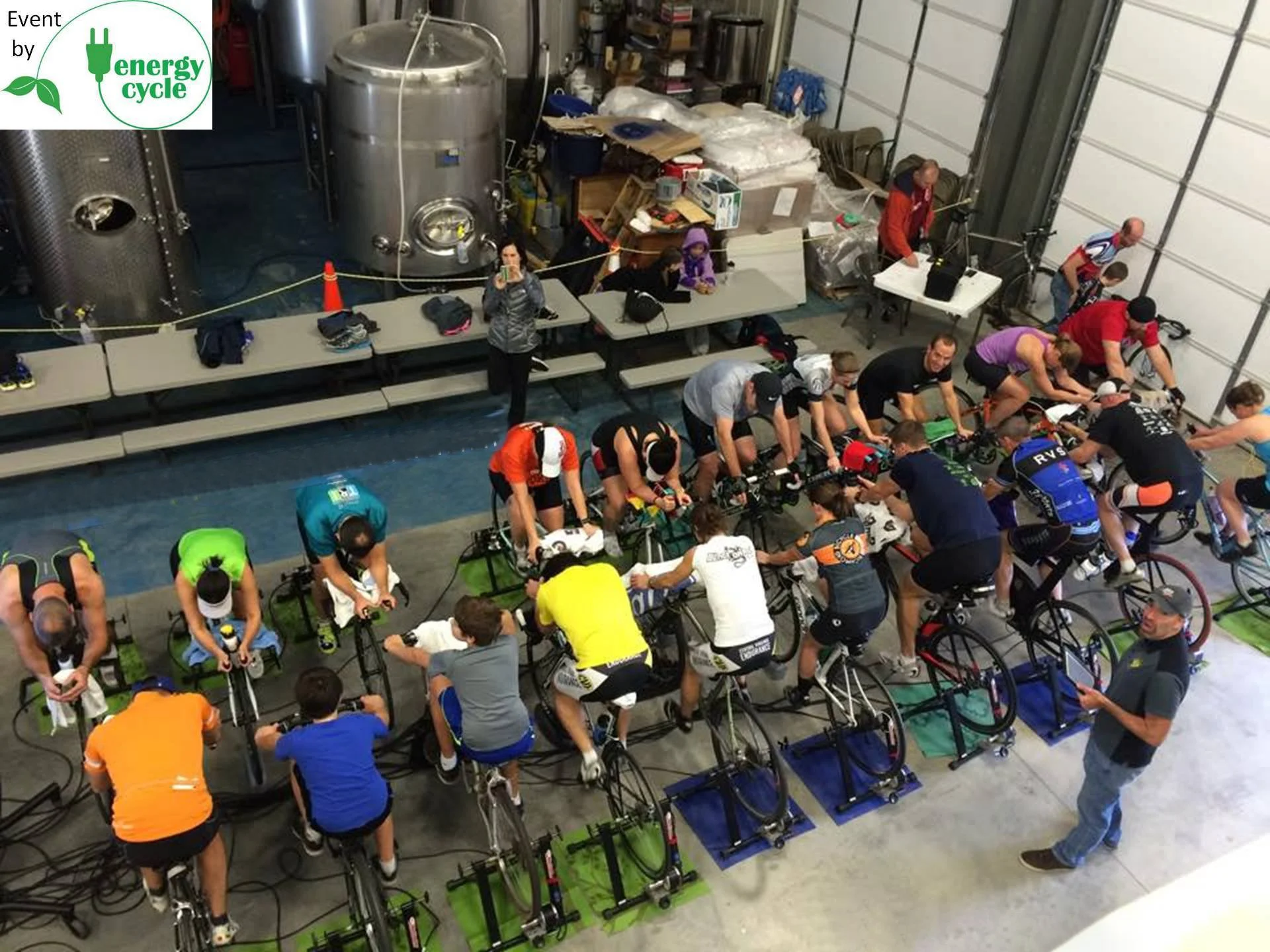Indoor cycling class with numerous participants on stationary bikes, some instructors, and a person holding a clipboard in a warehouse-like setting with large brewing equipment.