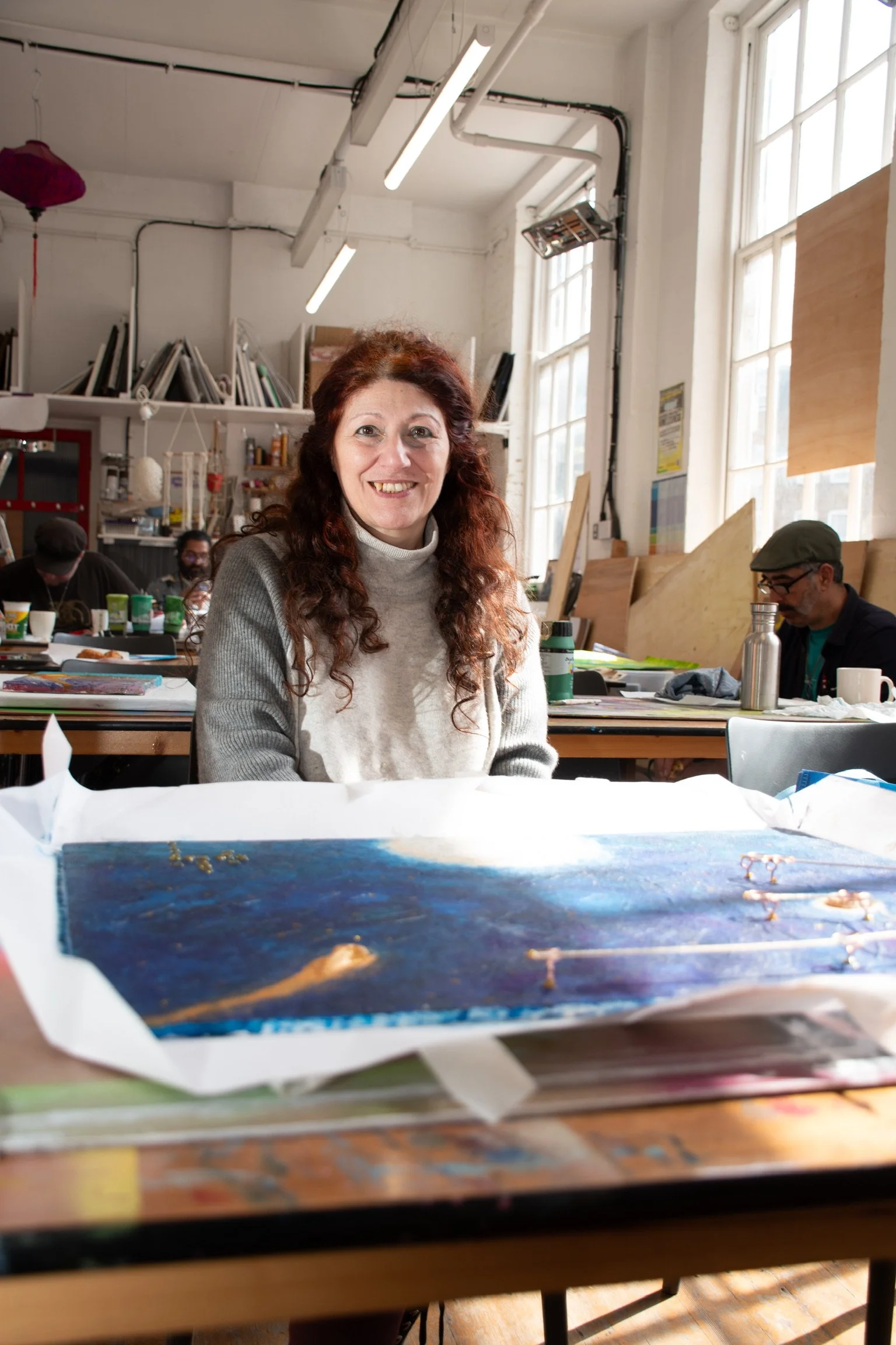 Smiling woman sitting in an art studio with a painting on the table.