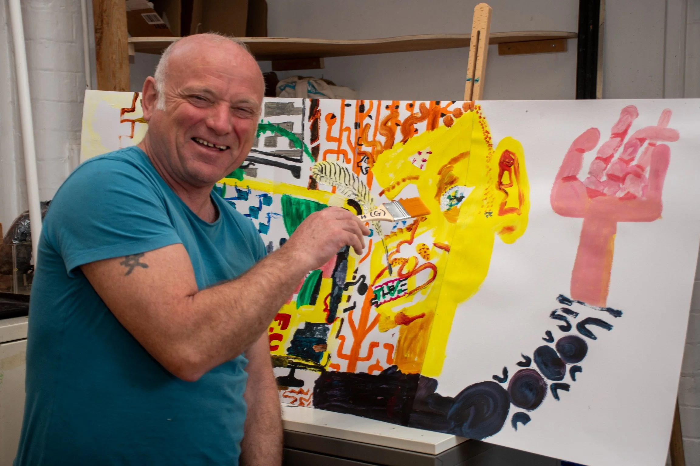 Person in a blue shirt smiling and holding a brush next to an abstract painting with vibrant colors and various patterns.