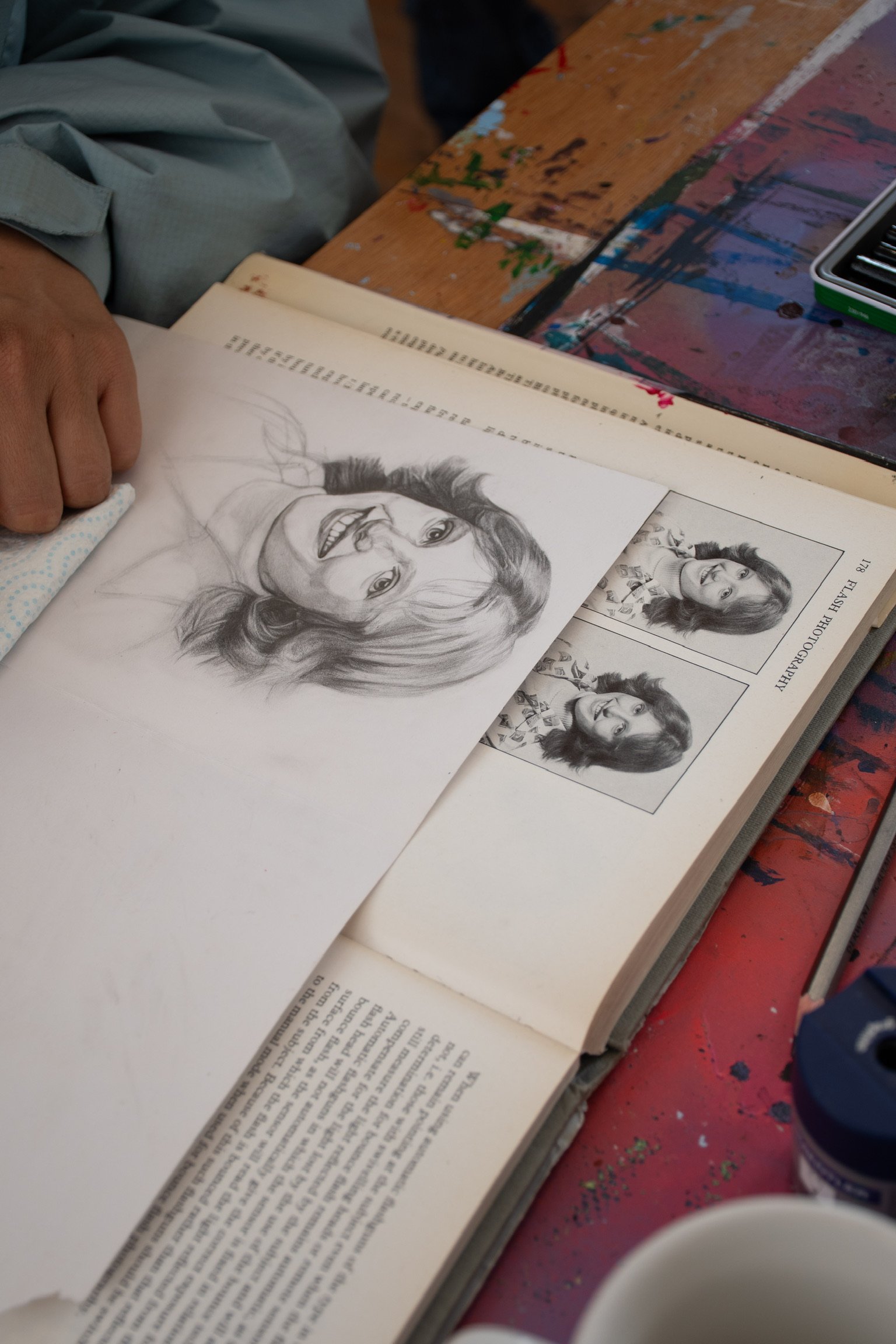 Person drawing a portrait on paper resting on an open book with photographs of a woman, on a paint-splattered desk.