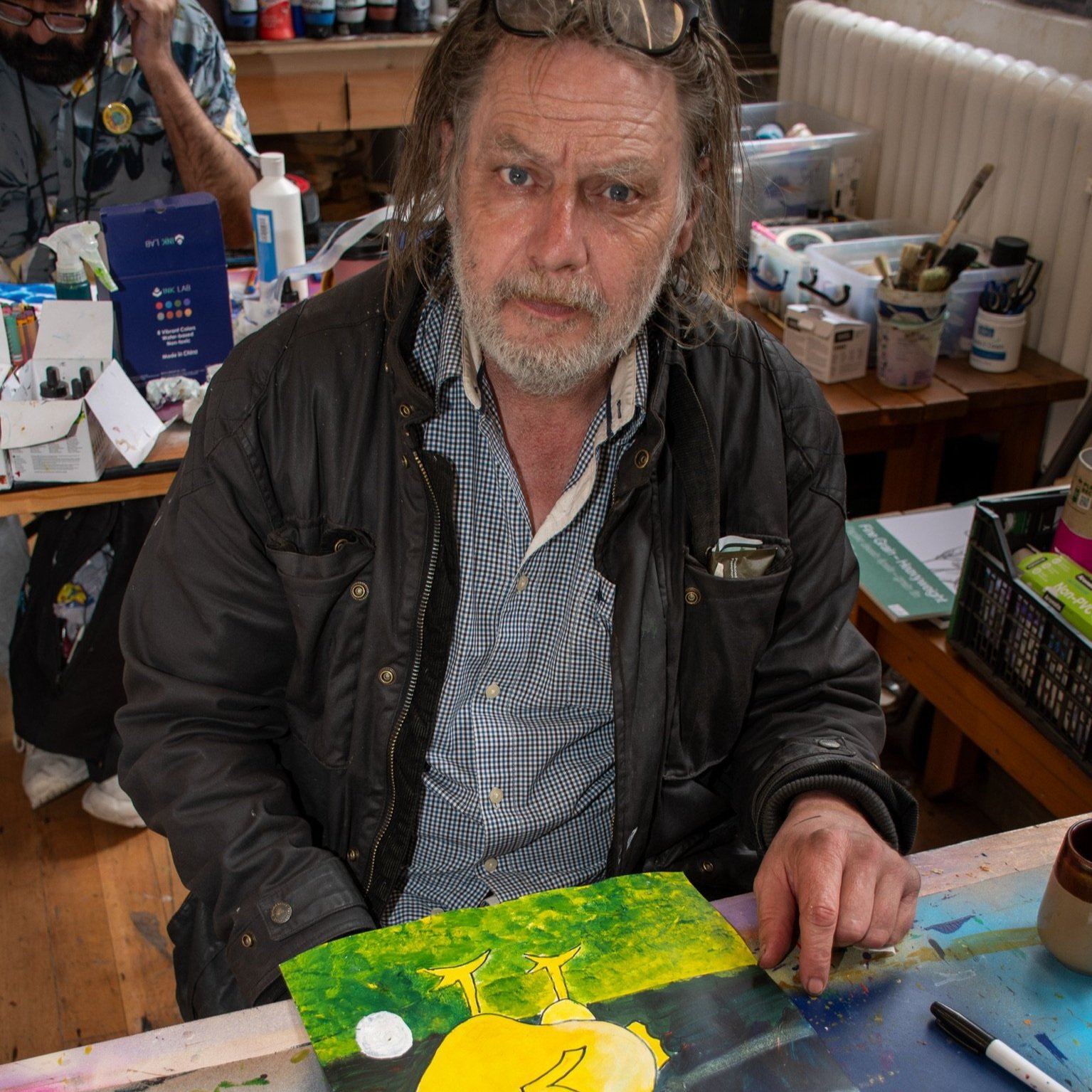 Man with long hair and glasses sitting at a table, pointing at a colorful painting on the table. Art supplies and brushes are visible around him. Another person is in the background, also engaged in art activities.