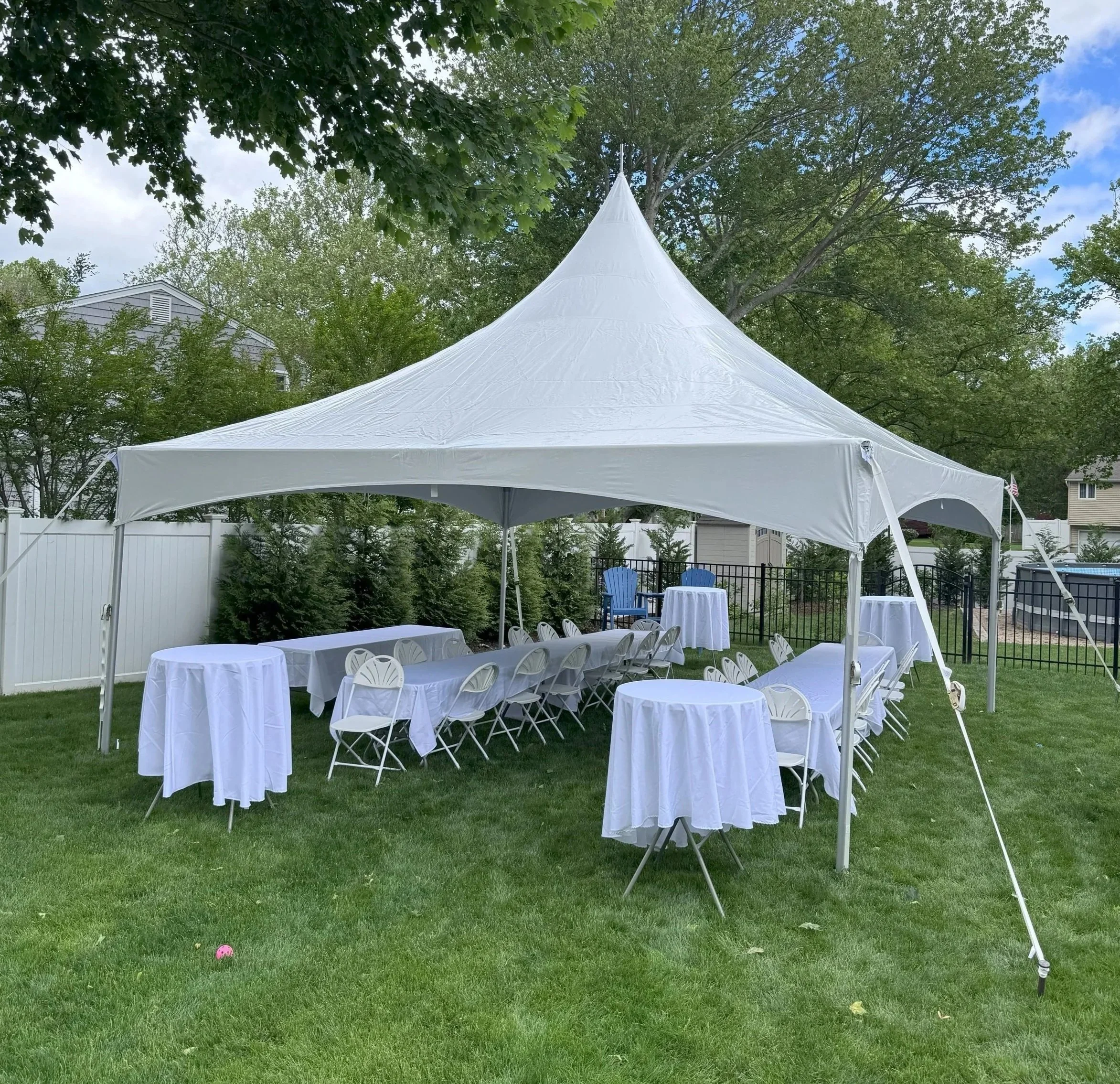 White outdoor event tent set up on a grassy lawn with tables and chairs covered in white tablecloths, surrounded by trees and houses.